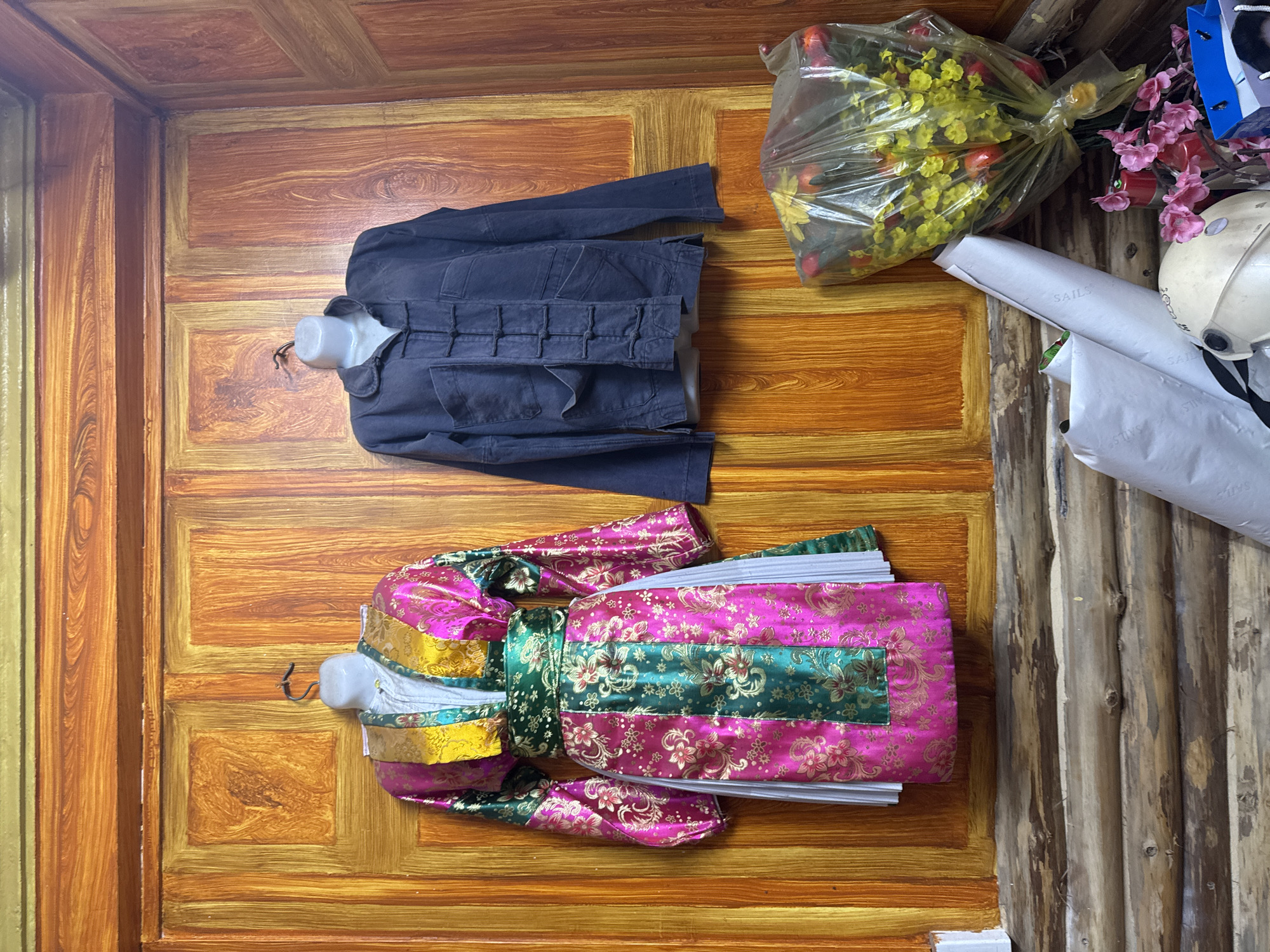 The image, likely taken in Mèo Vạc Commune, Vietnam, shows a scene with several objects placed on a wooden surface that appears to be a wall or possibly a door.
A dark blue jacket is presented on a white, partial mannequin. Below it, there's a colorful garment, possibly a traditional dress or robe, also displayed on a partial mannequin. This item features a vibrant combination of pink, green, and gold fabrics with an intricate floral pattern.
To the upper right, a plastic bag contains yellow and red flowers. Adjacent to this are other items: a white helmet, documents or rolled paper labeled "SAILS", pink decorative flowers, and a blue shopping bag.
The background consists of a wooden surface that varies from the painted wall behind the clothing to the raw, unfinished wood.