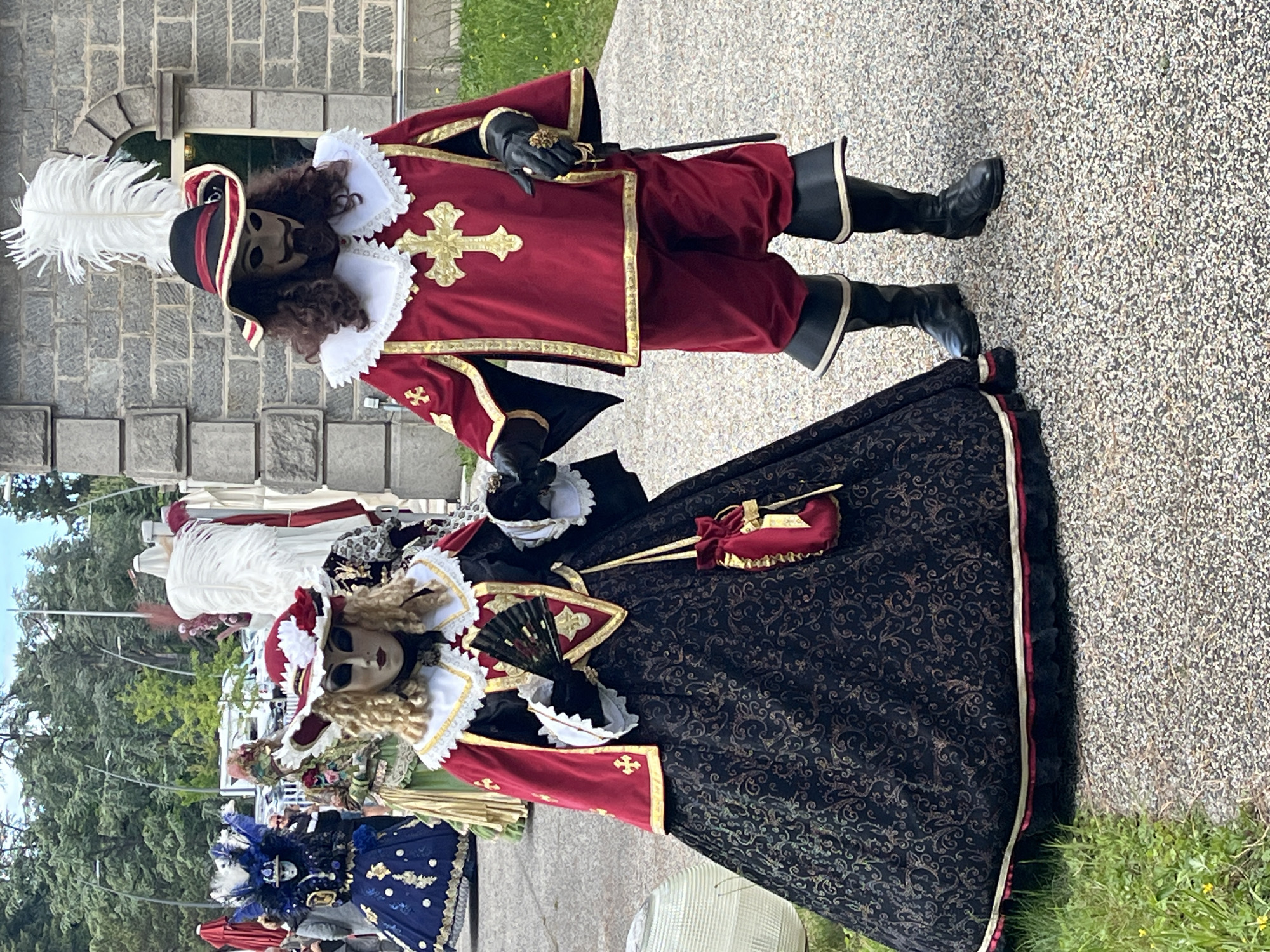 Two individuals in elaborate historical costumes stand on a gray stone surface, likely a walkway. The costumes evoke a period style, possibly 17th or 18th century, with elements like lace collars, rich fabrics, and decorative crosses.
One costume is primarily red with gold trim and features a masked character wearing a feathered hat and black boots. The other costume incorporates a dark, patterned fabric with gold and red accents and a long, elaborate skirt. The person is also wearing a mask and holds a fan.
In the background, other costumed figures can be seen, indicating an event or festival. Trees, a building, and overcast sky suggest an outdoor setting, likely in Saint-Galmier, France. The presence of other people in costumes, umbrellas, and other details add context to the scene.