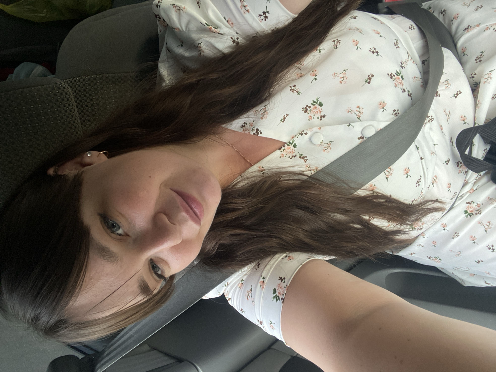 This is a selfie of a woman in a car, likely Votkinsk, Russia. She is wearing a white floral print dress and a grey seatbelt. The woman has long, dark brown hair and is looking at the camera with a slight smile. The lighting suggests it is daytime, and the interior of the car appears to be dark. The background shows a car seat, which is dark grey and textured. The image is taken from a high angle, with the woman's face filling a significant portion of the frame. The seatbelt is diagonally across her chest, and part of her arm is visible on the right side of the image.