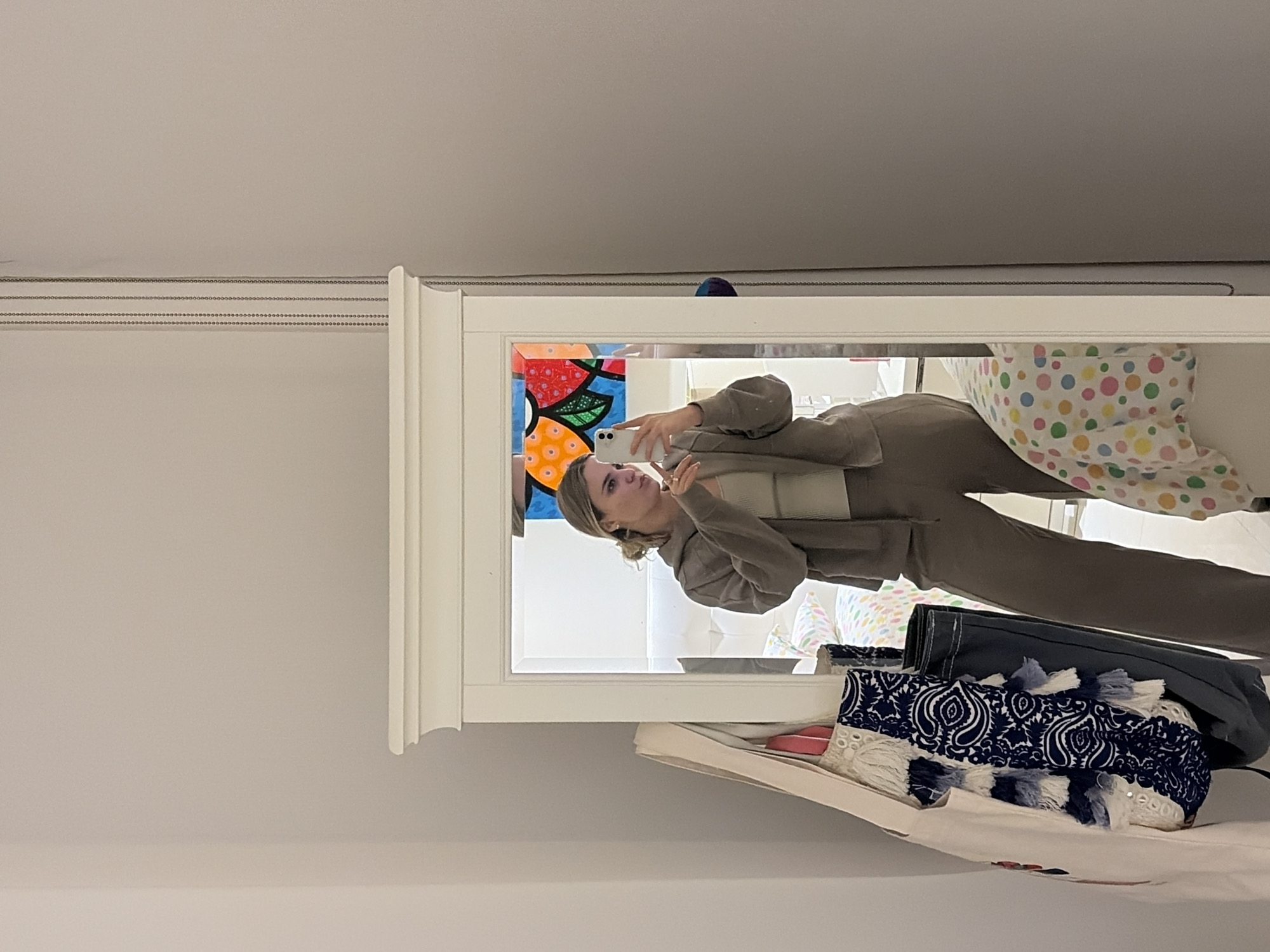 A woman is taking a photo of herself in a mirror. She is wearing a beige suit. The mirror is mounted on a wall, with a painting to the left. In front of the mirror are some items, including a handbag with a blue and white patterned scarf on top. The location is indoors, possibly in Key Biscayne, United States of America.