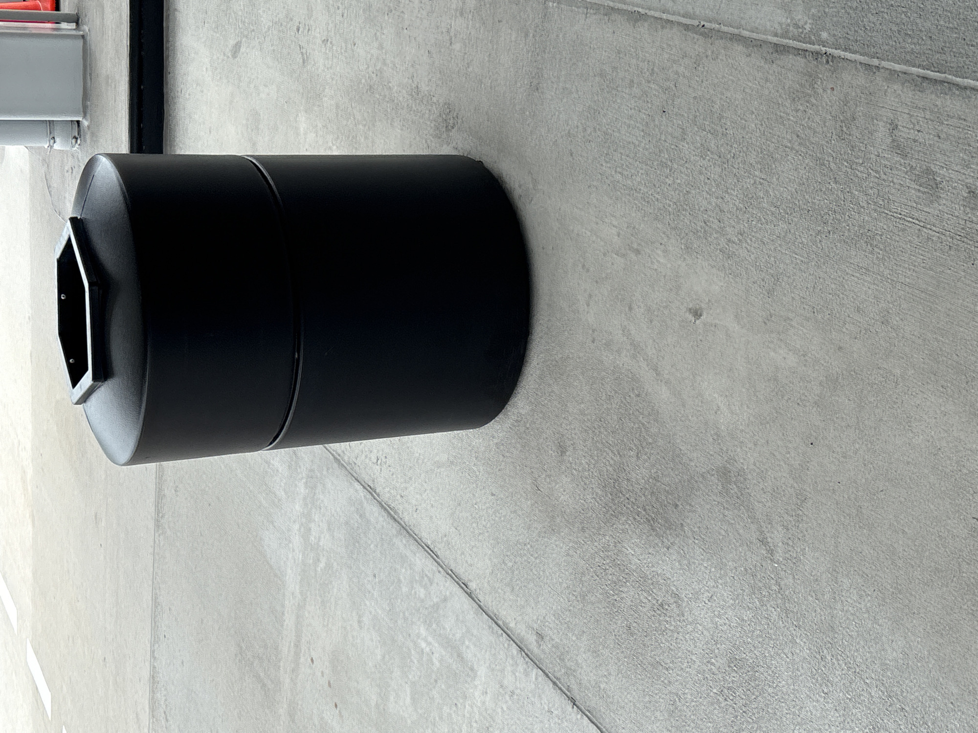 A black, cylindrical trash receptacle with a hexagonal opening on top sits on a gray concrete surface. The receptacle appears to be made of plastic or metal and has a smooth, matte finish. The concrete surface shows some expansion joints and is somewhat textured. In the background, there is a glimpse of a light-colored structure and possibly some orange traffic cones. The scene is outdoors and appears to be in an urban or public area, possibly a park or a commercial district. The image is taken from a slightly elevated perspective, looking down at the trash receptacle.