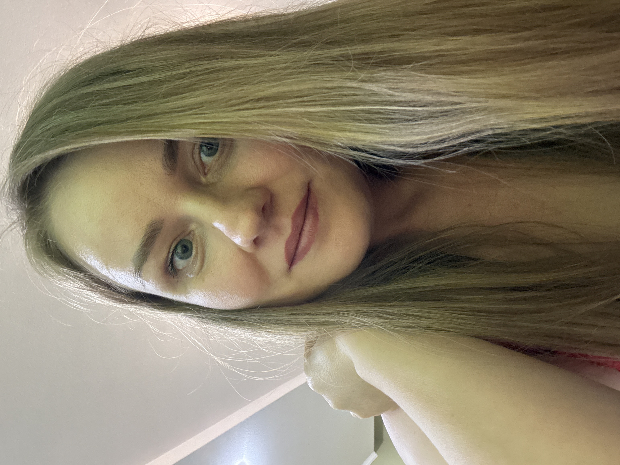 An indoor portrait captures a young adult woman with long, straight, blonde hair, looking directly at the camera with a gentle, inviting smile. Her hair, a blend of blonde and light brown tones, cascades over her shoulders and partially across her forehead. Her eyes are a striking blue-green, complementing her fair complexion. She has natural-looking eyebrows and a soft, warm expression with her lips slightly parted, revealing a subtle, natural lip color or light lipstick.
The woman is positioned as if reclining or lying down, with the camera looking down at her. Her right arm or hand is visible, resting under her chin or supporting her head, though largely obscured by her hair and the framing of the shot. A hint of red fabric, possibly from her clothing, is visible on the lower right side of the frame. The background is a plain, light color, likely a wall or ceiling, creating a clean and uncluttered setting. A subtle bright light source or reflection is visible in the bottom left corner. The overall lighting is soft and natural, highlighting her features and giving her skin a smooth appearance. The image conveys a sense of calm and approachability. There is no visible text. The location context provided for this image is Chaikovsky, Russia.