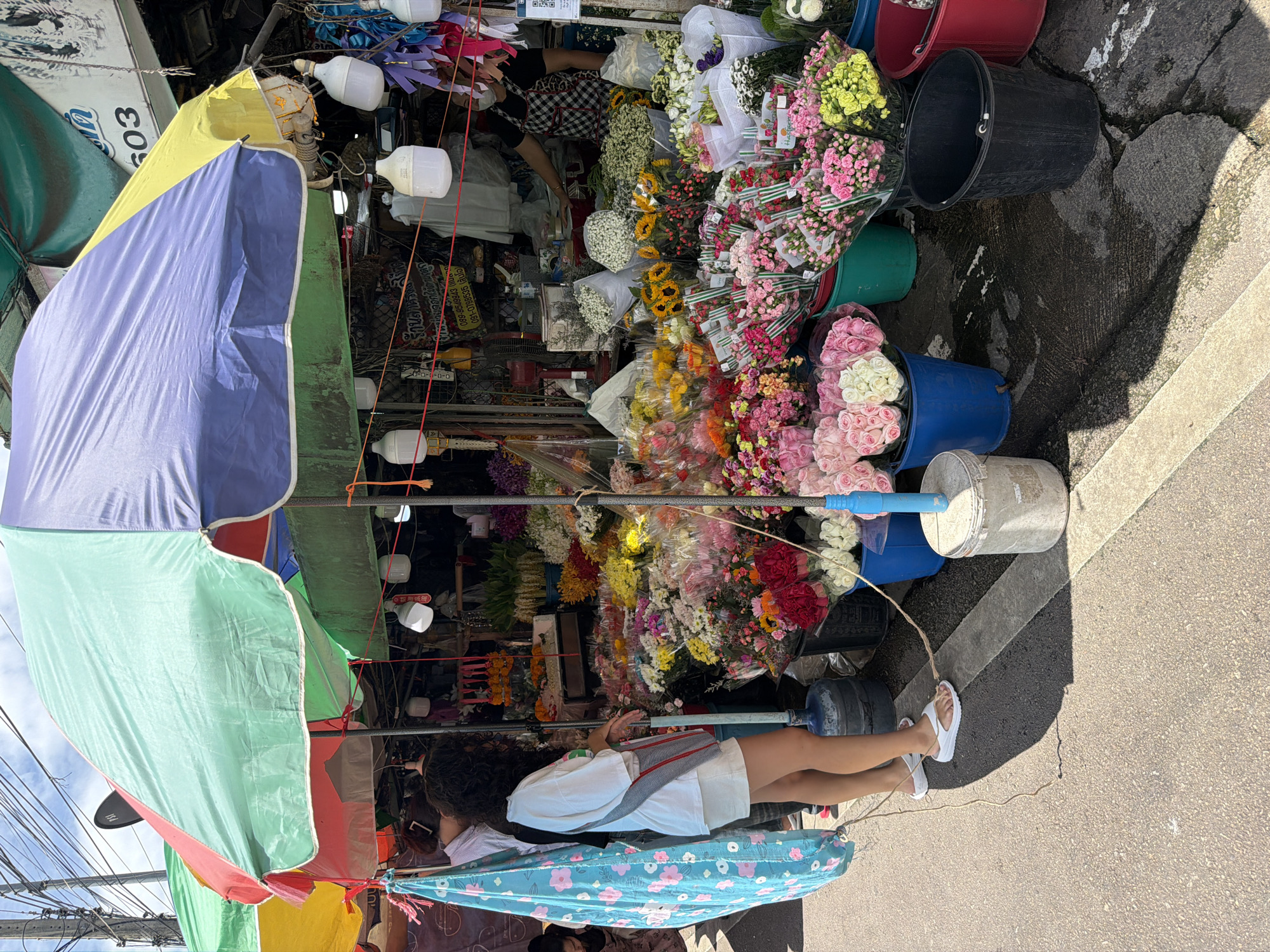 A street-level outdoor flower market is depicted, likely in Chiang Mai City Municipality, Thailand, under a large, multi-colored umbrella. The foreground shows a portion of a woman with curly hair, wearing a white t-shirt and white shorts, with white flip-flops. She is standing in front of a large display of flowers. Her left arm is slightly bent, and her hands are near her waist. A blue cloth with pink floral patterns hangs to her left. Behind her, a variety of colorful flowers are arranged in buckets and on shelves. These include roses, sunflowers, and other assorted blooms in shades of pink, yellow, white, red, and orange. Several large plastic buckets, some containing flowers and others empty, are visible. In the background, a stall is set up with more flowers and what appears to be merchandise. A person wearing a black and white checkered apron is visible behind the counter, partially obscured by the flowers. Electrical wires and lighting fixtures are suspended above the stall, suggesting it is a functional marketplace. The ground is paved with asphalt and concrete, with distinct lines suggesting a pedestrian walkway or parking area. Shadows are cast on the ground from the umbrella and surrounding structures. The overall impression is a vibrant and busy flower market on a sunny day.