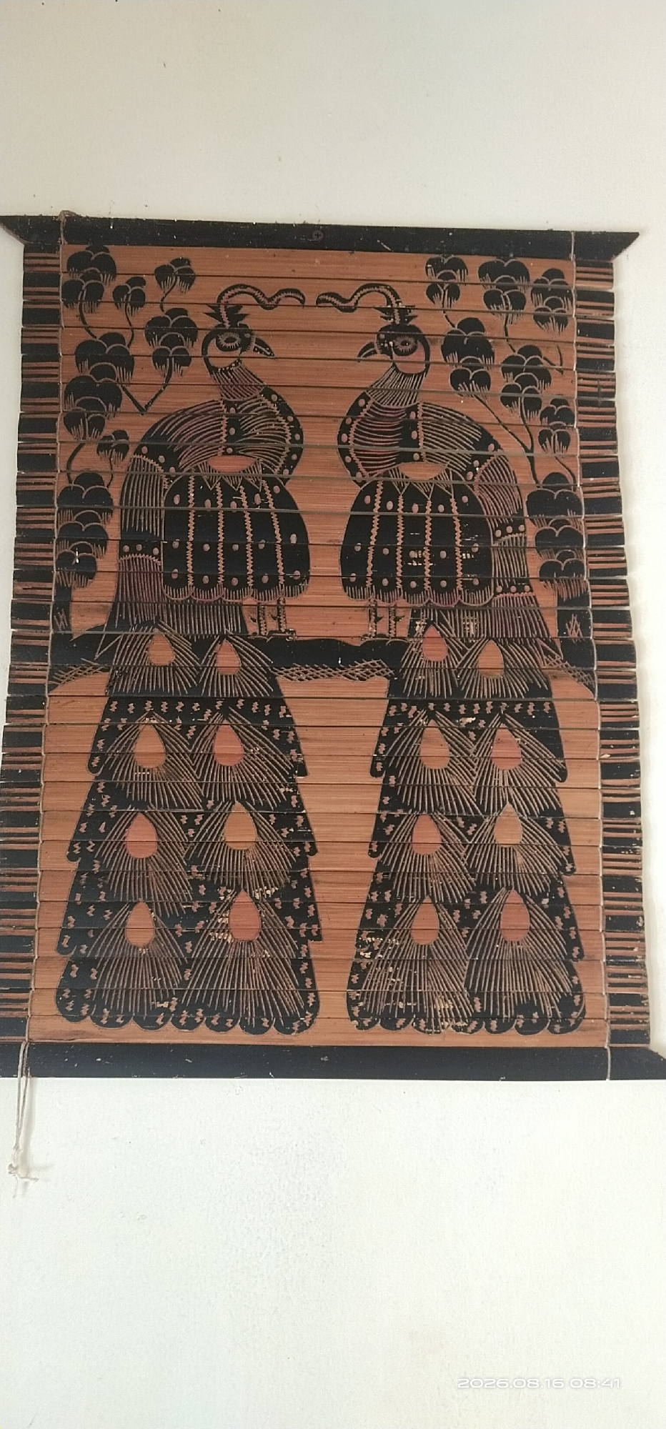 The image shows a close-up of a bamboo blind with a black and orange design featuring two stylized peacocks. The peacocks are positioned symmetrically, facing each other, with their tails fanned out. The design appears to be painted or wood-burned onto the bamboo slats. The blind is hanging against a white wall. There is a thin white string hanging from the left side of the blind. The overall impression is of a decorative home accessory. No specific location in Muka Haji, Indonesia is identifiable from the image.
