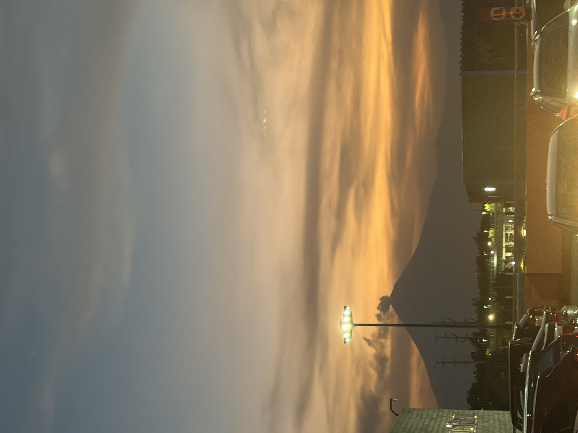 The image captures a dramatic outdoor scene at dusk or twilight, likely in Puebla, Mexico, featuring a volcano and an urban landscape. The sky dominates the upper and left parts of the frame, showcasing a striking blend of colors, with warm golden and orange hues on the right transitioning to cooler blue-gray tones on the left. Streaky clouds are illuminated by this ambient light, creating dynamic patterns.
In the mid-ground, the dark, imposing silhouette of a large mountain or volcano, consistent with Popocatépetl, stands out against the vibrant sky. A small plume or cloud is visible near its summit, suggesting volcanic activity. Below the mountain, an urban environment is visible with several dark buildings, some displaying illuminated windows or signs. On one building, indistinct white shapes resembling "99" or "GG" are visible on a dark banner.
Artificial lighting plays a significant role in the scene, with a prominent multi-lamp streetlight brightly illuminating the lower-middle section. Numerous smaller lights dot the background, and the tops and partial views of several parked cars are visible in the foreground and to the right, some with illuminated headlights or taillights. Utility poles and power lines crisscross parts of the mid-ground. The overall atmosphere conveys the serene beauty of an evening sky contrasted with the formidable presence of the volcano and the subtle glow of urban life.