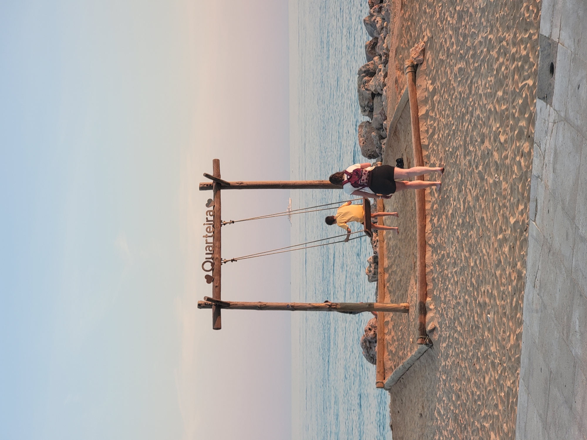 This is a full-frame, eye-level, vertical shot taken at sunset on a sandy beach in Quarteira, Portugal. The image features a wooden swing set with "QUARTEIRA" spelled out in metal letters above it. A young person in a yellow shirt is on the swing, facing away from the camera. A woman with a backpack and dark shorts is standing on the sand to the right of the swing, also facing away. The sea is calm and blue behind them, with a boat visible in the distance. The sand is golden and textured, with a paved walkway in the foreground. Rocks are scattered along the shoreline. The sky is a soft gradient of blue and pink.