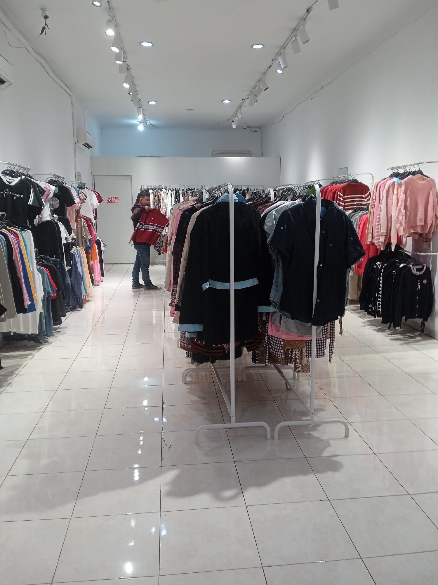 The image depicts the interior of a clothing store located in Semarang, Indonesia. The store has white tiled flooring and white walls, with a track lighting system installed on the ceiling. Racks of clothing are displayed on both sides of the aisle, organized by color and style. In the background, a young man wearing a dark hooded jacket and blue jeans is standing and examining a red garment with a patterned design. He is holding the garment with both hands. A white door is visible behind him. The overall impression is that of a typical retail environment for apparel.