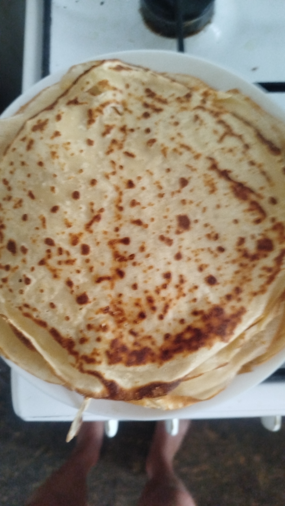 A close-up, top-down shot displays a stack of thin, golden-brown crepes or pancakes on a white plate. The crepes exhibit a slightly uneven surface with darker brown, irregularly shaped spots scattered across them, indicating they have been cooked on a griddle. The stack of crepes is positioned on a white surface that appears to be a stovetop, with the edge of a black burner visible in the upper background. Below the plate, the lower legs and feet of an unseen person are visible, suggesting they are either standing or sitting very close to the cooking area. The overall environment appears to be domestic, likely a kitchen. The image was taken in Kostiantynivka, Ukraine.