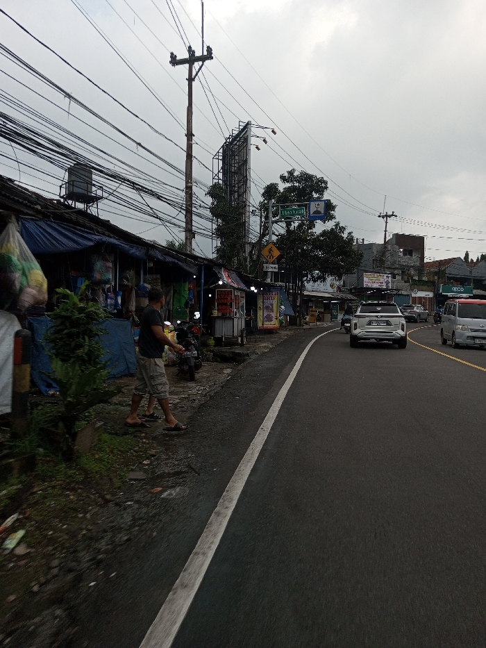 This image depicts an active street scene under an overcast, gray sky. Visual evidence from signage, specifically the text "BADRUM" and "SELALU PAS", indicates the location is an Indonesian-speaking region, contradicting the provided location of Dawathang_Dorjibi_ Kashingtsawa, Bhutan.
On the left side of the frame, a row of makeshift roadside stalls or small shops are visible. These structures feature blue tarpaulin coverings and corrugated metal roofs. A black water tank is present on the roof of the leftmost stall. Merchandise, including items in clear plastic bags, hangs from the stalls or is displayed on rudimentary shelves. Adjacent to these stalls, a male individual is standing on the unpaved ground, wearing a dark short-sleeved shirt, light-colored shorts, and dark sandals. He is positioned near a dark-colored scooter parked on the dirt. Small green plants are present along the immediate roadside. A prominent wooden utility pole heavily laden with a dense network of black electrical and communication cables dominates the upper left and central portions of the scene.
The road itself is a dark asphalt surface, curving gently to the right as it extends into the background. A solid white line delineates the edge of the road from the unpaved shoulder. Further down the road, a double yellow line marks the center. Several vehicles are in motion: a light-colored SUV with roof racks is visible heading away from the camera in the lane furthest to the right, followed by another vehicle. A light-colored van is visible in the opposing lane, approaching the camera.
In the mid-distance and background on the right side of the road, various buildings of different heights are visible. Several trees with green foliage are interspersed. A tall, empty metal framework for a billboard stands behind the main utility pole. Road signs are also present, including a blue rectangular sign with white text (possibly "BADRUM"), another blue sign with a mosque symbol, and a yellow diamond-shaped pedestrian crossing warning sign. The overall lighting is diffused, consistent with an overcast day.