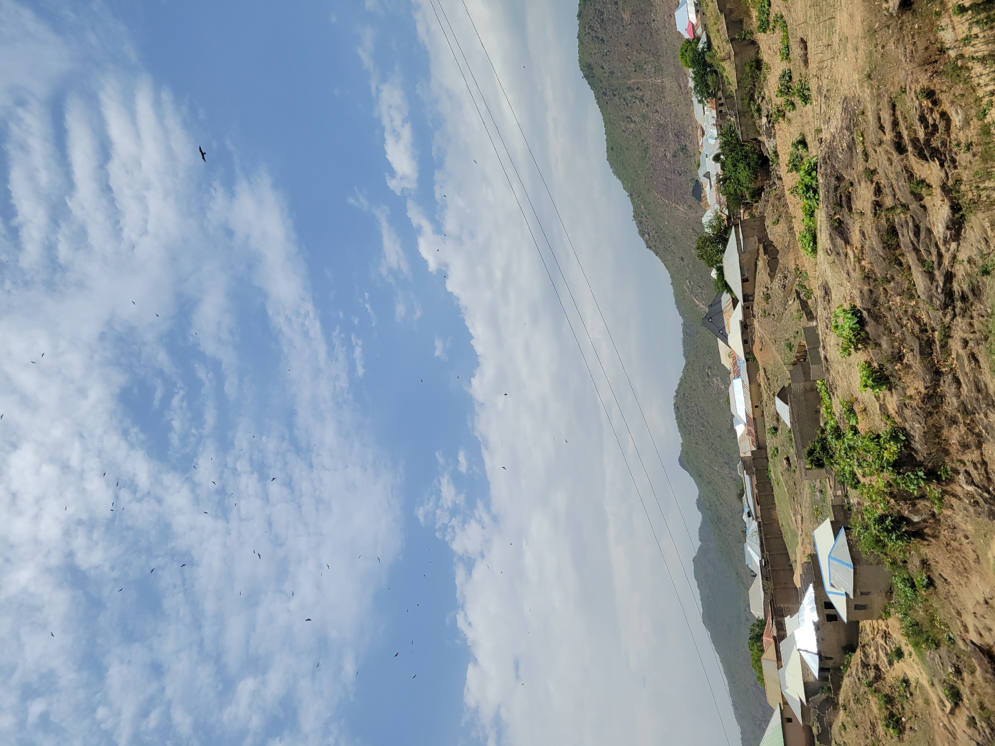 The image shows a village situated on a hillside in Tilde, Nigeria. The houses have corrugated iron roofs, some painted in bright colors. The hillside is steep and rocky, with sparse vegetation. In the sky, a flock of birds fly against a backdrop of clouds and blue sky. High-tension powerlines are strung across the landscape.