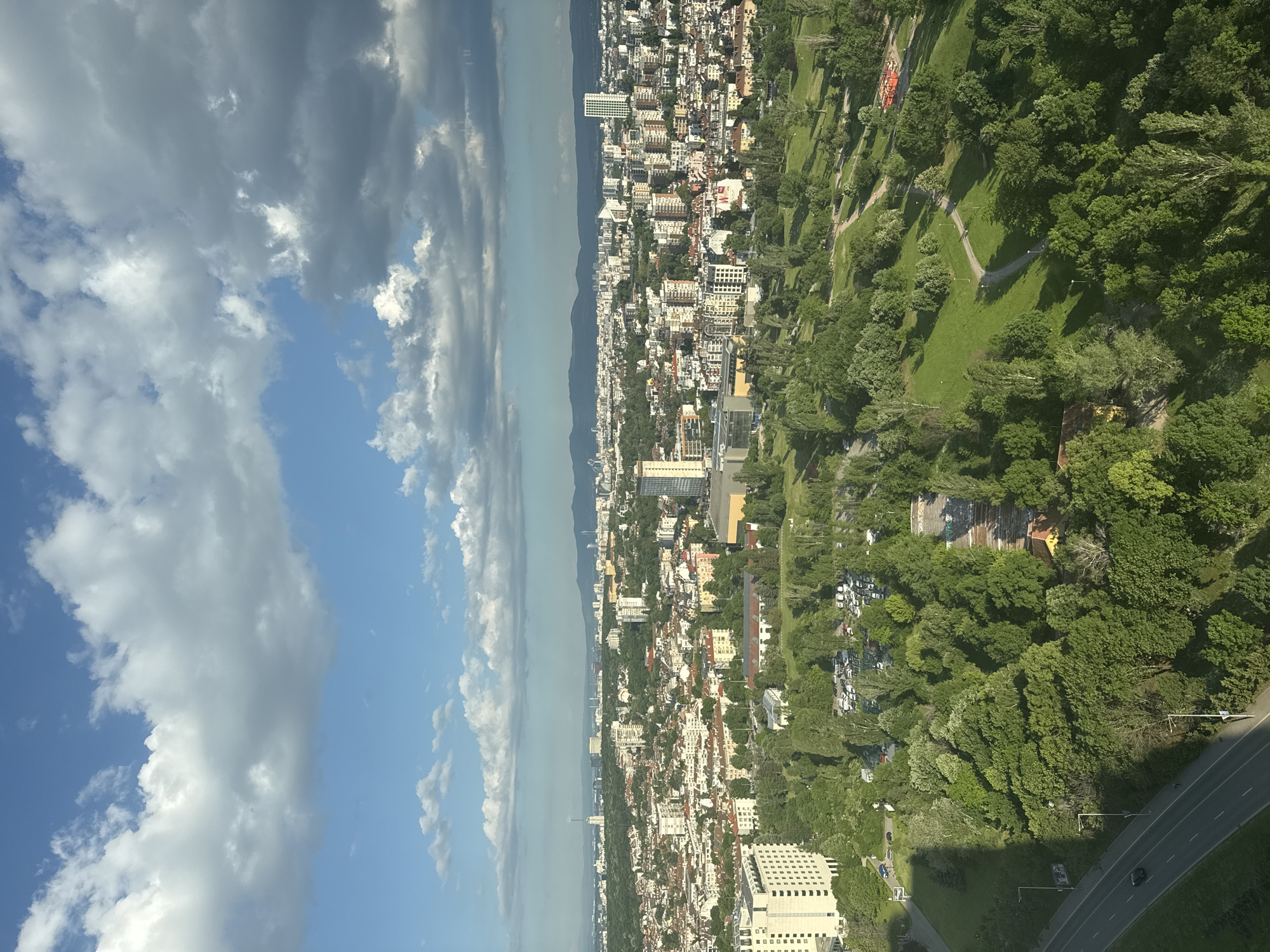 This is an aerial view of Sofia, Bulgaria, capturing a cityscape interspersed with green spaces. The image shows a densely packed urban area with predominantly white and light-colored buildings. The backdrop features a mountain range. A park with walking paths and trees is seen to the right of the buildings. The sky is blue with scattered cumulus clouds. A highway runs along the bottom right.