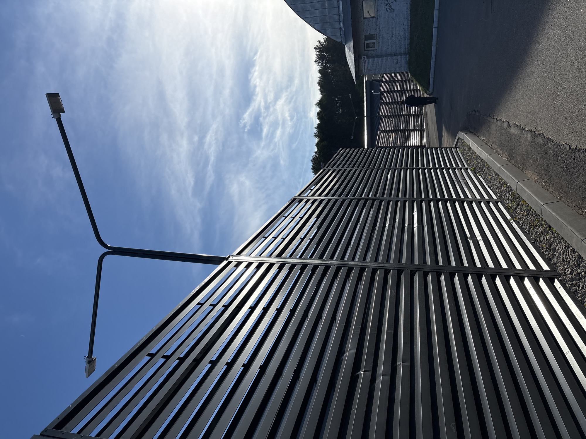The image depicts an outdoor scene on a bright, partly cloudy day, likely in the city of Izhevsk, Russia. A person, silhouetted and walking away from the camera, is visible in the middle ground on a paved pathway. To the left, a tall, dark, slatted metal fence dominates the foreground and extends into the background. Above the fence, a street lamp with two fixtures extends diagonally across the frame against a blue sky with wispy white clouds. In the background, to the right of the fence and further down the pathway, is a low-rise brick building with a light-colored, possibly painted, facade. There are some trees visible in the distance, beyond the fence and buildings. The ground beside the fence is covered in dark gravel. The scene appears to be an industrial or commercial area.
