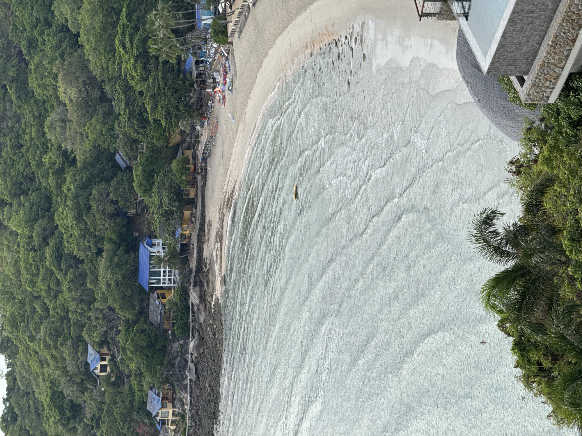 A high-angle, vertical shot captures a tropical beach scene. The foreground is dominated by the turquoise, rippling ocean, with small waves lapping at the shore. A solitary figure can be seen swimming in the water. Beyond the water, a sandy beach stretches out, dotted with beach chairs and umbrellas, indicating a resort or hotel. Several structures with bright blue roofs are nestled into the lush green hillside that rises behind the beach, suggesting a small village or a collection of villas. Palm trees and dense foliage cover the hillside, creating a vibrant, natural backdrop. A modern structure with a stone facade and an infinity pool is visible in the lower right corner, overlooking the beach. The overall impression is one of a serene and picturesque coastal landscape.