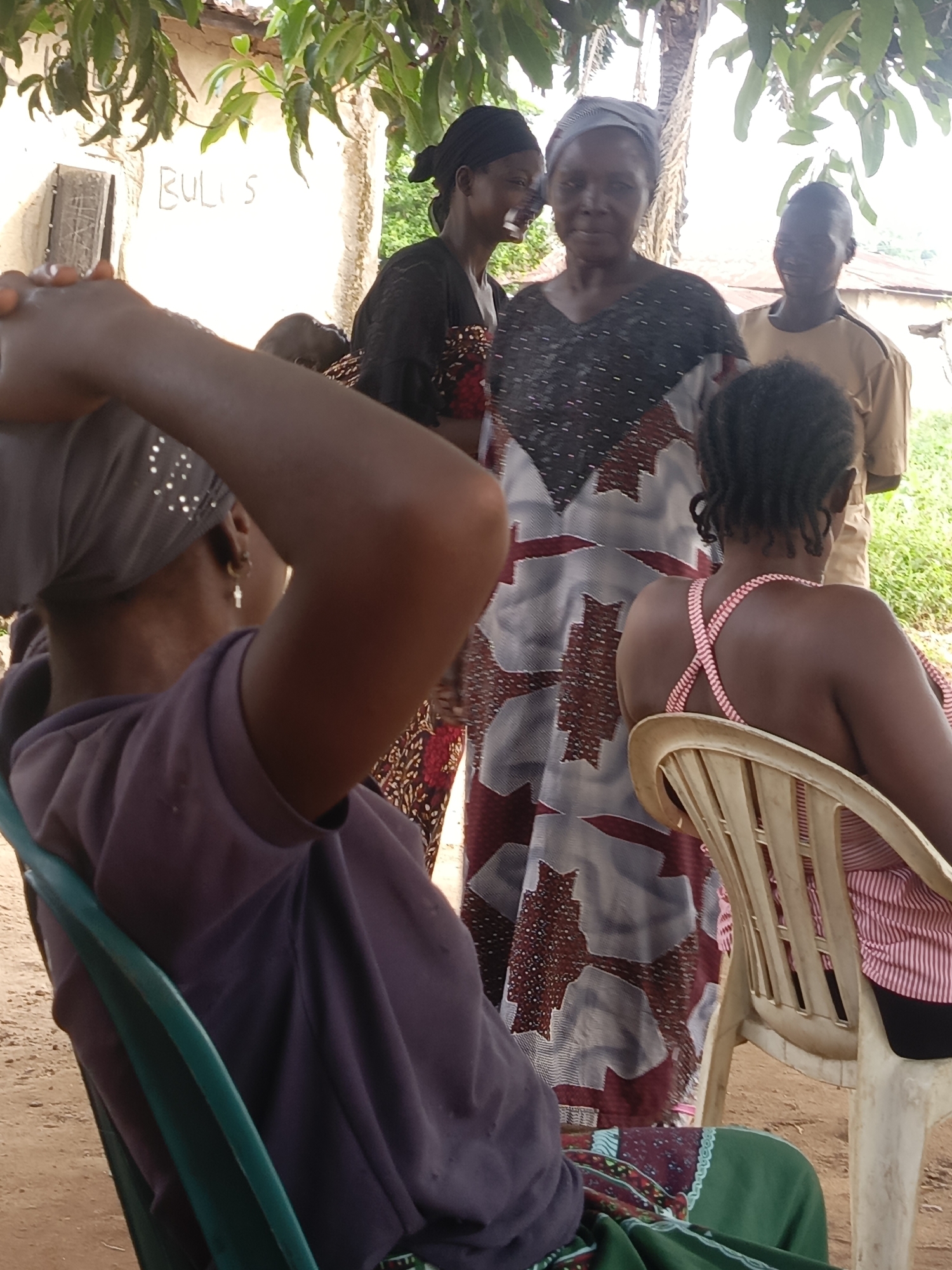 In Unguwar Zaria, Nigeria, several people are gathered outdoors. In the foreground, a person wearing a purple shirt and head covering is seated in a turquoise chair, with their back to the viewer. To the right, another individual in a pink and white striped top sits on a light beige chair, also facing away from the camera. Standing behind them is a woman in a gray and maroon patterned dress with a gray headscarf. To her left, a woman in a dark patterned dress seems to be speaking to her. Behind them, a person in a tan uniform is visible. A building with the word "BULI S" written on it is in the background. Overhanging the group is foliage from trees. The ground appears to be dirt.
