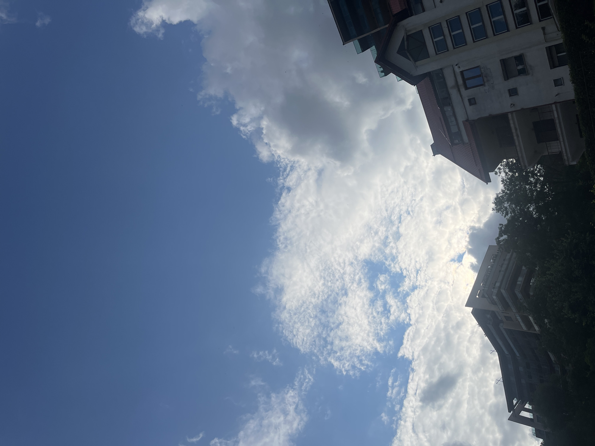 The image captures a view of buildings and the sky in Brasov, Romania. On the left, a modern, multi-story building with a distinctive angular design is visible, partially obscured by trees. To the right, a more traditional building with a red-tiled roof, balconies, and numerous windows stands out. The sky above is a mix of clear blue and scattered clouds, with sunlight filtering through. The scene appears to be a daytime shot, likely from a slightly elevated perspective looking upwards. No people or specific activities are discernible in the image.