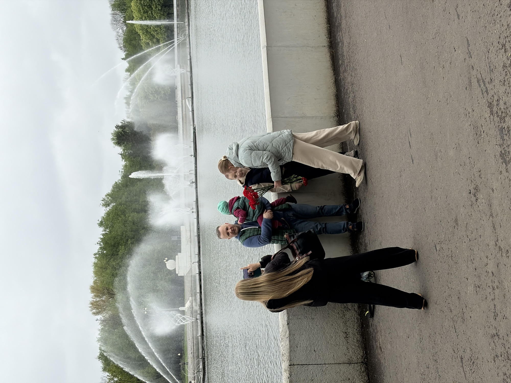 Here is a detailed description of the image:
**Scene Overview:** The photograph captures a family scene near a large water fountain in what appears to be an urban park. The fountain features multiple jets of water spraying high into the air.
**People:** There are several individuals in the scene:
* A woman is taking a photo of the other family members.
* A man is holding a young child.
* A woman with blonde hair and a light-colored jacket is standing next to the man.
* Another woman stands next to the woman in light jacket.
**Objects:**
* The water fountain with multiple spraying jets.
* Flowers are held by one of the individuals, specifically the child.
* A phone is being used to take a photograph.
**Actions & Interactions:**
* The woman is actively taking a photograph of the family.
* The family appears to be interacting with the fountain and posing for pictures.
**Environment:**
* The backdrop consists of trees and a somewhat overcast sky, suggesting a park-like setting.
* The location is near a concrete barrier or wall next to a pavement or sidewalk.