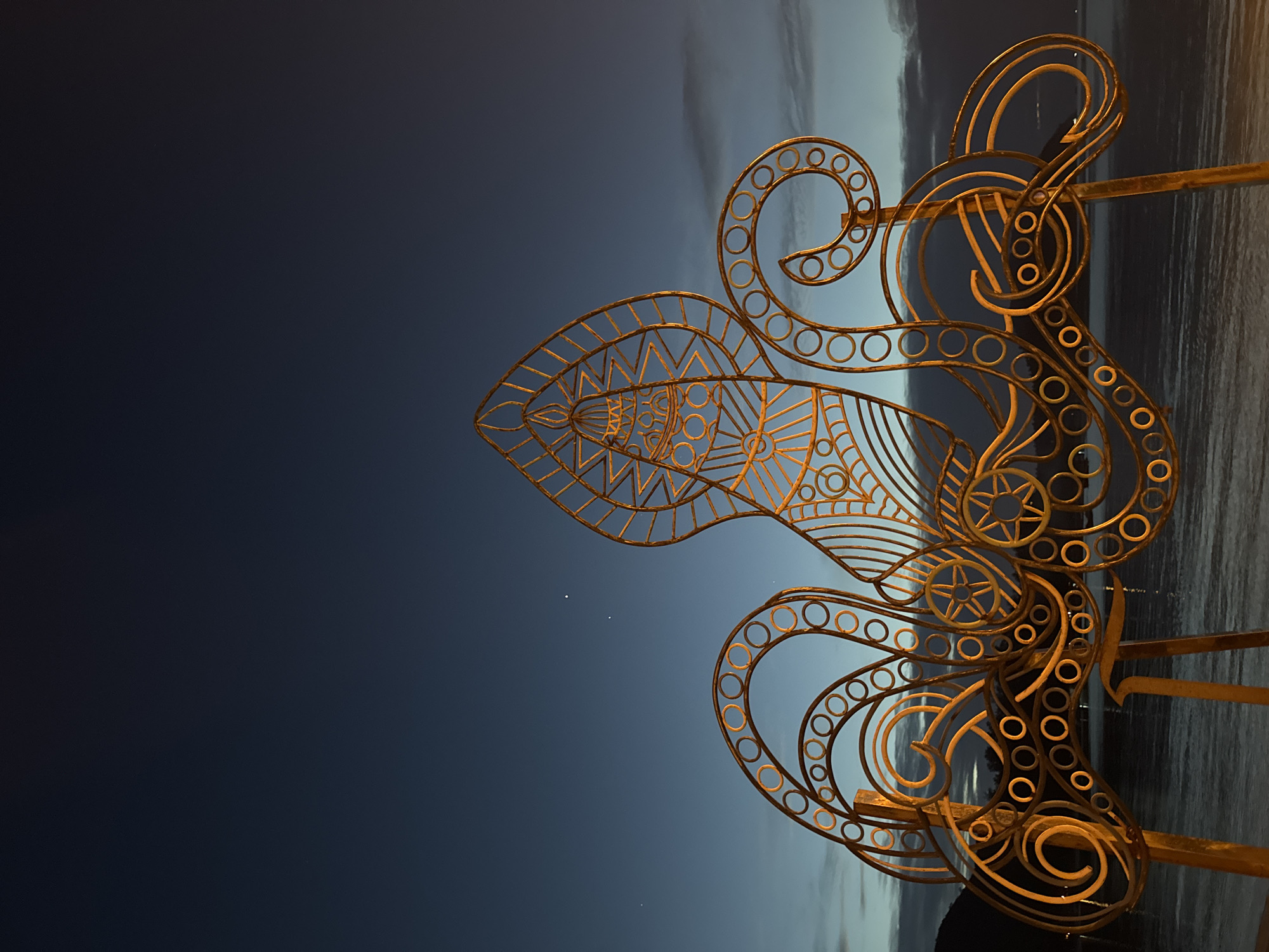 A large, golden, metal sculpture, resembling an octopus with intricate geometric patterns, is silhouetted against a twilight sky over a body of water. The sculpture features a central, elongated head with triangular designs and numerous swirling tentacles adorned with circular cutouts. The sky transitions from a deep navy blue at the top to lighter hues of blue and grey near the horizon, with wisps of clouds. A faint glow emanates from the sculpture, suggesting it is illuminated. In the distance, the dark outline of a landmass is visible. The water reflects the dim light, creating a calm surface. There are three small, bright points of light visible in the sky, possibly stars or planets. The sculpture appears to be located outdoors, near a waterfront. This scene is located in Koh Phangan, Thailand.