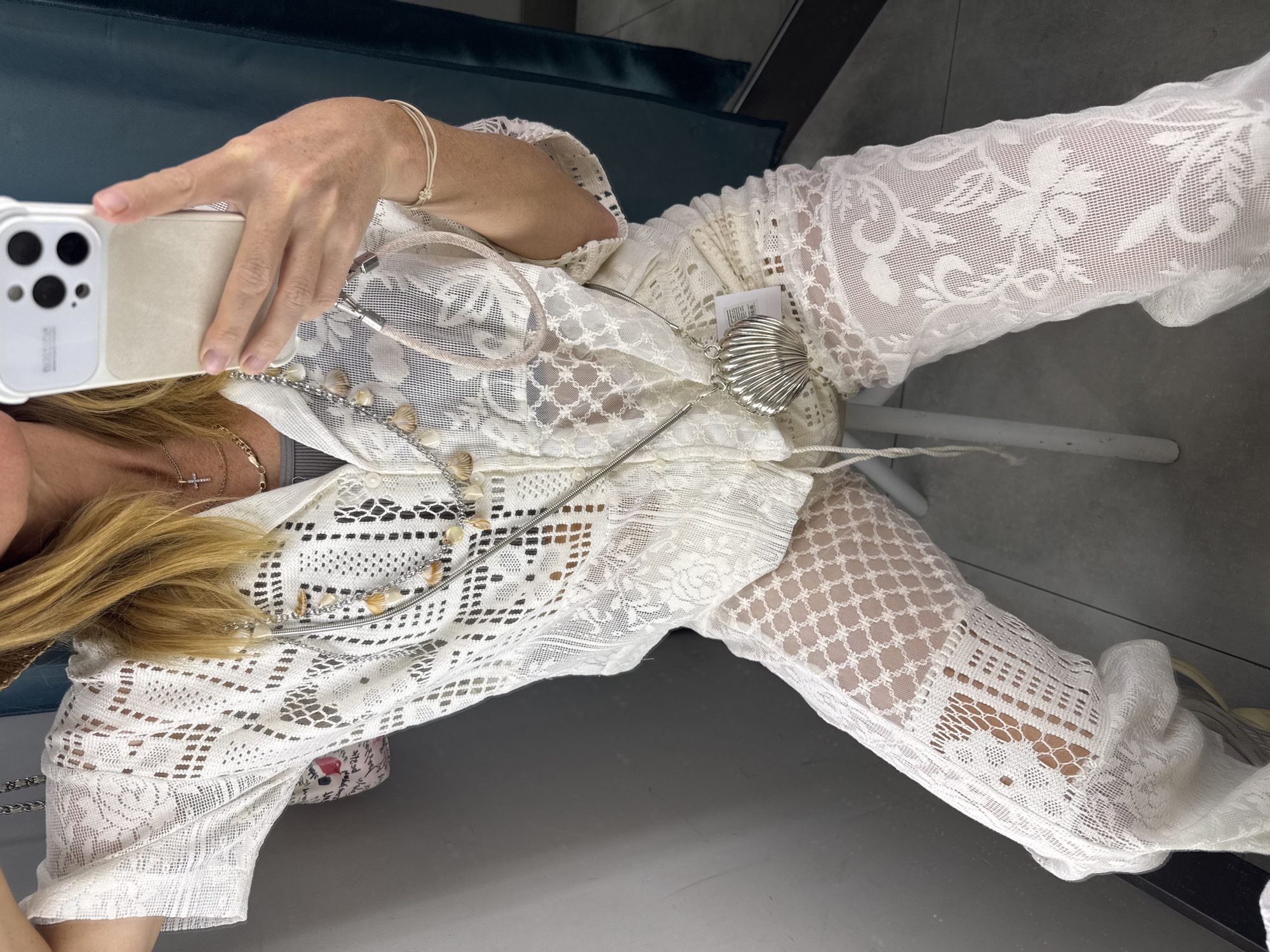 A woman takes a selfie in a fitting room. She is wearing a white lace outfit and several necklaces. She is holding a smartphone in a white case. The fitting room has a blue velvet bench and a gray floor. The location is unknown, but not specified in the prompt to be Saint Petersburg, Russia.