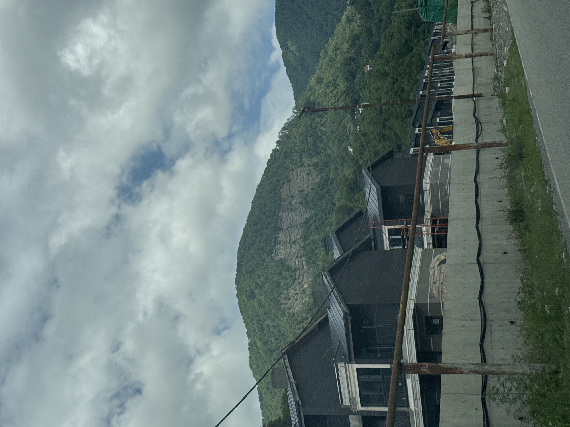 The image captures a view of a construction site with modern houses under construction in the foreground, set against a backdrop of lush green mountains and a cloudy sky. The houses are dark in color with metallic roofs and feature balconies. A concrete wall with rusted metal railings runs along the front of the property, with exposed construction materials and equipment visible behind it, including a yellow excavator. In the distance, the mountains are covered with dense vegetation, and a few small structures are visible on the slopes. The scene appears to be a residential development in a mountainous area. The location is identified as Plastunka, Russia.