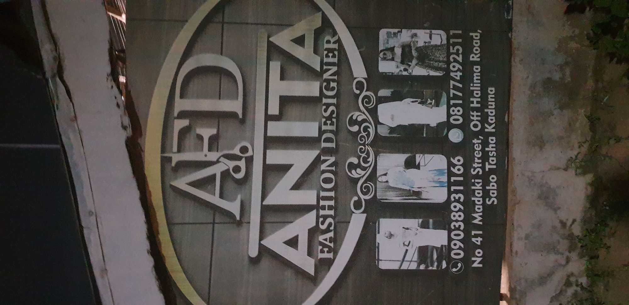 The image shows a sign for "AFD ANITA FASHION DESIGNER". The sign is oval-shaped with a metallic border. The letters "AFD" are at the top, with a pair of scissors in place of the "F". Below this, the name "ANITA" is written in larger font, and below that, "FASHION DESIGNER". At the bottom of the sign, there are three small square images of women posing in outfits, and contact information. The phone numbers listed are 09038931166 and 08177492511. The address is "No 41 Madaki Street, Off Halima Road, Sabo Tasha Kaduna". The sign appears to be mounted outdoors, possibly on a building. The background of the sign is a textured, dark material, possibly wood or stone. The overall presentation suggests a business that designs and creates clothing.