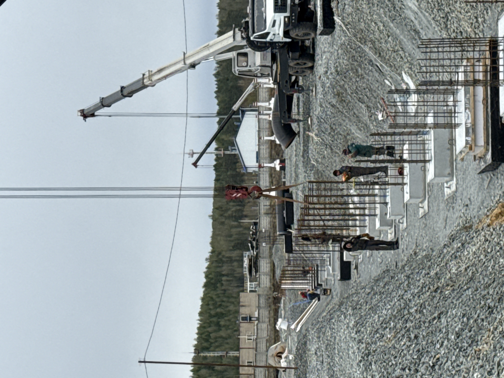 The image shows a construction site near Leninsk, Russia. A large white crane truck is parked on a bed of gray gravel. Several workers are visible near concrete and rebar structures, presumably working on foundations or supports. In the background is a line of small, modular buildings and a forested area. Overhead power lines are visible against an overcast sky.