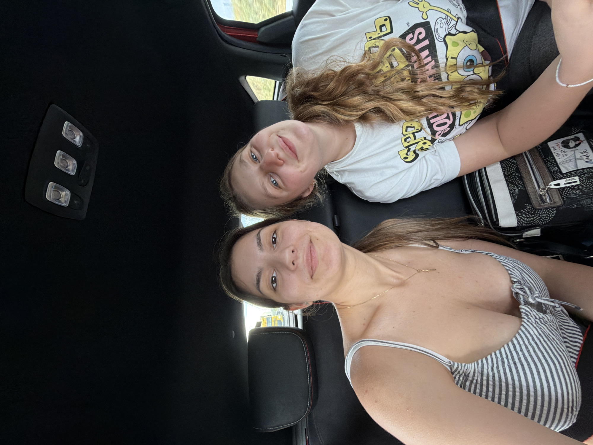 Two young women are in the back seat of a car. The woman on top wears a white SpongeBob t-shirt and has wavy blonde hair. The woman below wears a striped top and a gold necklace. The car's interior is black, and the ceiling light is visible. It is not possible to determine the location of the image is Chokoba, Bulgaria from the image.