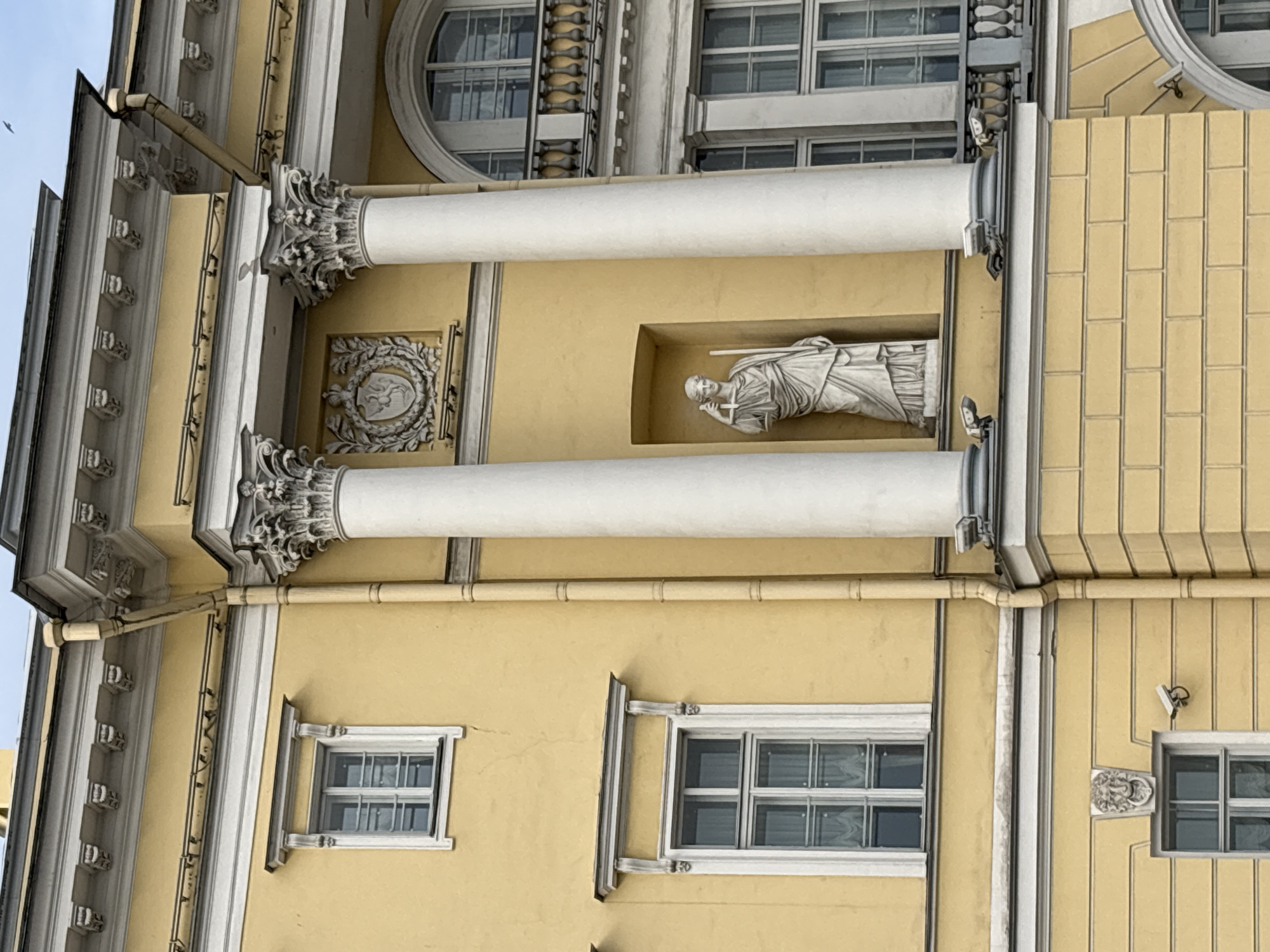 This is a close-up, low-angle exterior, eye-level shot of a grand, yellow classical-style building in Saint Petersburg, Russia. The building features ornate architectural details, including fluted Corinthian columns, decorative moldings, and arched windows with balconies. A white marble statue of a robed figure holding a staff and a cross is set in a niche between two columns. The sky is overcast.