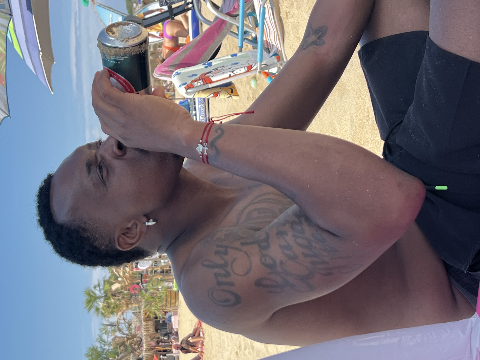 A man is drinking from a can on a beach. He has tattoos on his left arm and a bracelet on his right wrist. He is wearing black shorts. In the background, there are palm trees, beach umbrellas, and other people. The scene appears to be in Sozopol, Bulgaria, given the presence of a beer can with Bulgarian text that reads "BEER BREMEN DOBA" which is a Bulgarian beer brand and this beer is produced in Bulgaria.