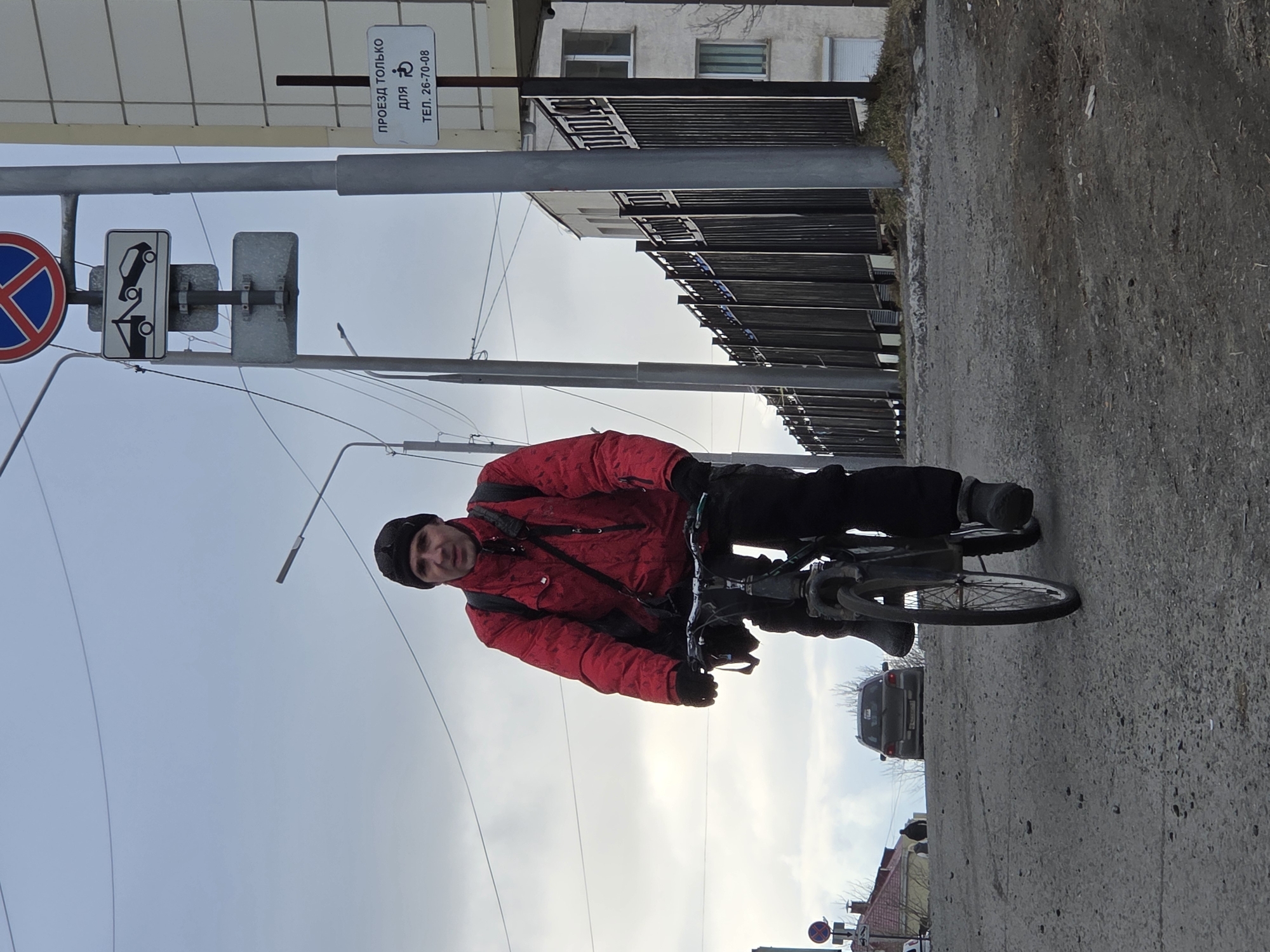 A man is riding a bicycle on a paved road in an urban environment. He is wearing a red jacket, black pants, and a black beanie. The background includes a building with black metal railings, power lines, street signs, and a grey sedan. The street signs feature a "no entry" symbol, a sign allowing access only for certain vehicles, and a sign indicating access for disabled people.