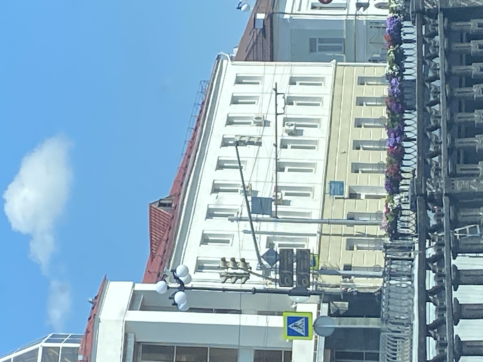 The image shows a view of buildings and a street in Kazan, Russia, on a clear, sunny day with a few clouds in the blue sky. The primary focus is on a white, multi-story building with a red roof, featuring numerous windows. To the left, a portion of another white building with a glass-topped structure is visible. A street light with multiple globes stands on the left side of the frame, next to a pedestrian crossing sign and traffic signals. Further down the street, another building with a light blue facade and a darker red roof is partially visible. In the foreground, a decorative metal railing runs along what appears to be a walkway or bridge, with planters filled with colorful flowers placed on top. The overall scene depicts an urban environment with architectural elements and floral decorations.