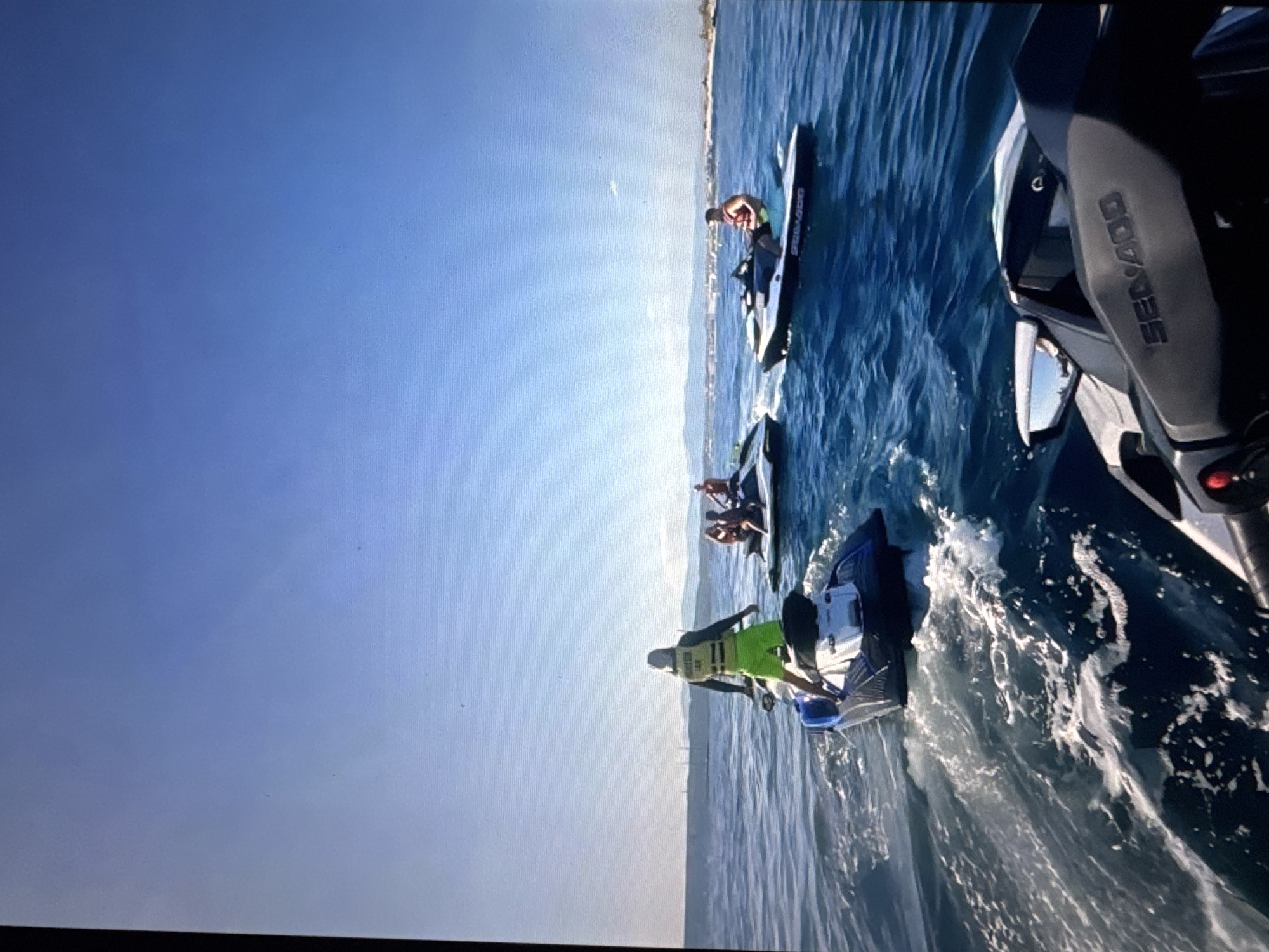 In a bright, sunlit scene, four individuals are enjoying a recreational activity on jet skis in a body of water, likely the Baltic Sea, with a distant coastline visible. The sky is clear and blue, meeting the calm, blue water. The perspective is from a jet ski in the foreground, looking out towards the others.
One person, wearing a yellow life vest and green shorts, stands on a blue and white jet ski in the middle ground, facing away from the camera and towards the sea. To their right, two people are seated on a white jet ski, one wearing a life vest. Further to the right, another person is seated on a black and white jet ski, wearing a red life vest and looking to their left. The jet skis are leaving white wakes in the water, indicating movement.
In the background, a hazy coastline with buildings and hills can be seen under the bright sky. The water is a deep blue, with gentle waves. The overall impression is one of leisure and outdoor activity on a clear day. The location is unlikely to be Tallinn, Estonia, as the landscape and type of water activity do not align with typical representations of Tallinn's coastline or popular recreational spots within the city.