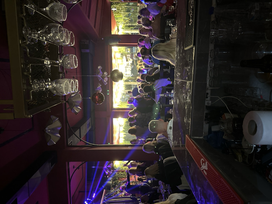This image captures the interior of a lively bar or club, likely situated in Peristeri, Greece, during the evening. The perspective is from inside, looking towards a large expanse of windows that offer a glimpse of an outdoor setting with trees and what appears to be a street or walkway.
Numerous patrons are visible throughout the venue, predominantly seated at tables or along a prominent bar counter. They appear to be engaged in conversation, socializing, and enjoying drinks in a relaxed atmosphere. The casual to semi-formal attire of the individuals suggests an evening out.
In the foreground to the left, a metal rack displays several clear wine glasses hanging upside down, reflecting the ambient light. Below this, decorative elements such as wavy silver metal forms and spherical hanging lights—one metallic and one reddish—add to the venue's aesthetic. The interior is illuminated with a blend of purple and blue accent lights, including distinct blue light beams, indicative of a dynamic or entertainment-focused environment. Scattered green dots across the midground suggest the presence of a disco ball or laser lighting effects.
The right side of the image features a dark, polished bar counter, well-stocked with various bottles and bar implements. A roll of white paper is visible on the counter, beside a red, textured bar mat that bears a partially obscured white logo or text. Outside the window, a sign with some legible white text, including the word "KING," can be faintly seen. The interplay of bright indoor lighting against the darkening exterior confirms an evening or nighttime setting. The overall impression is that of a bustling and modern establishment, offering a vibrant space for social interaction and entertainment.