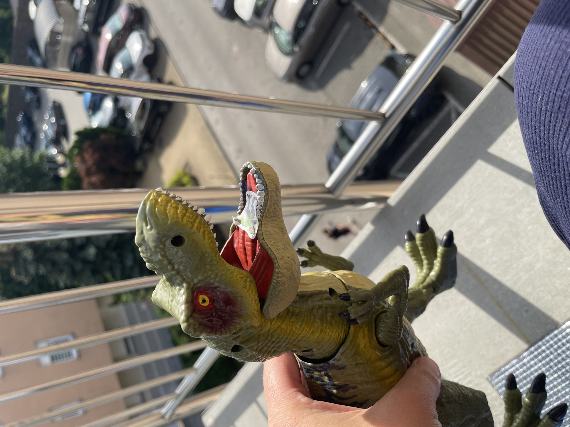A person's hand holds a green toy dinosaur with its mouth open, revealing a small, colorful sticker inside. The dinosaur is positioned in the foreground, with its head and upper body visible. In the background, a blurred urban environment is seen, including buildings, trees, and parked cars. The scene appears to be outdoors, possibly on a balcony or a similar elevated structure, given the presence of a metal railing. The lighting suggests a sunny day.
