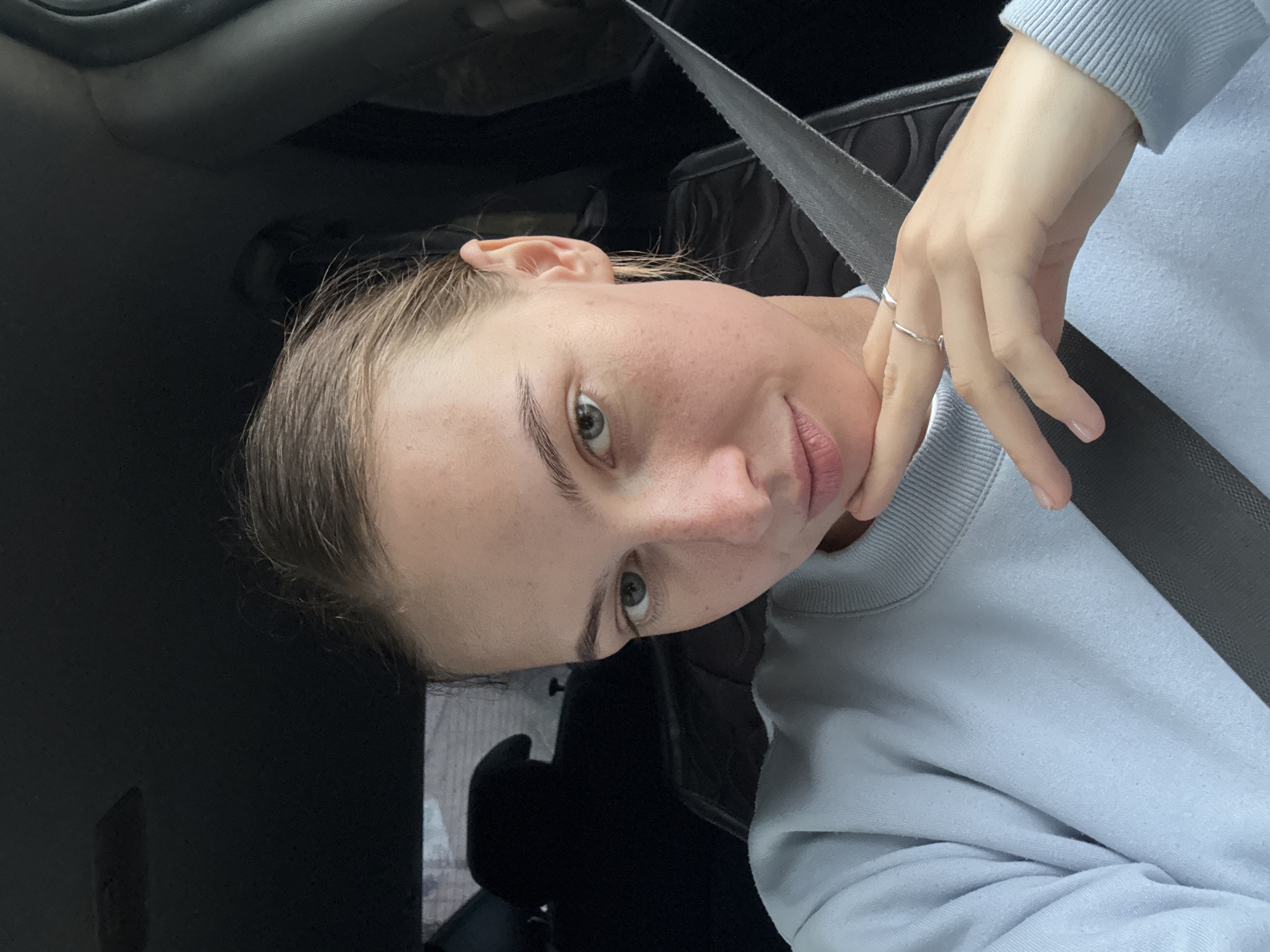 A young woman with fair skin and light brown hair is sitting in the driver's seat of a car. She is looking directly at the camera with her head tilted slightly and her left hand resting on her chin. She is wearing a light blue sweatshirt and a silver ring on her left index finger. The interior of the car is visible, including the seatbelt, part of the headrest, and the dashboard. The background appears to be a street scene, with buildings visible through the car window. It is daytime, and the lighting suggests an overcast sky.
