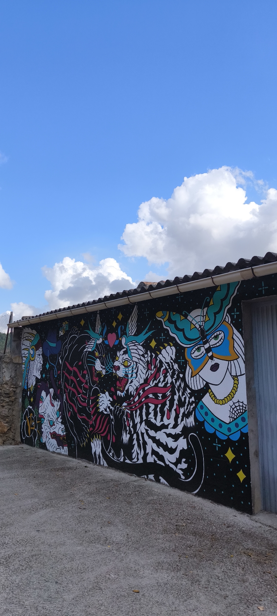 The image shows a mural painted on the exterior wall of a building in Fanzara, Spain. The mural features a complex design with several elements. On the left, there are depictions of faces and creatures, including one that resembles a Native American headdress. The central part of the mural is dominated by two tigers, one black with red stripes and the other white with black stripes, locked in a dynamic pose. Both tigers have fierce expressions and open mouths, showing their teeth. Above them, two small birds are facing each other, with a pink heart shape between them. To the right of the tigers, there is a woman's face adorned with a butterfly-like mask and a yellow necklace. The background of the mural is black, covered in small yellow star-like shapes and some blue cloud-like forms. The wall itself is dark, likely black, serving as a canvas for the vibrant graffiti. The building has a tiled roof with a gutter running along the top edge of the mural. The ground in front of the mural is paved with concrete. The sky above is a clear blue with scattered white clouds, suggesting it is daytime. The overall scene is an outdoor public space showcasing artistic expression through street art.