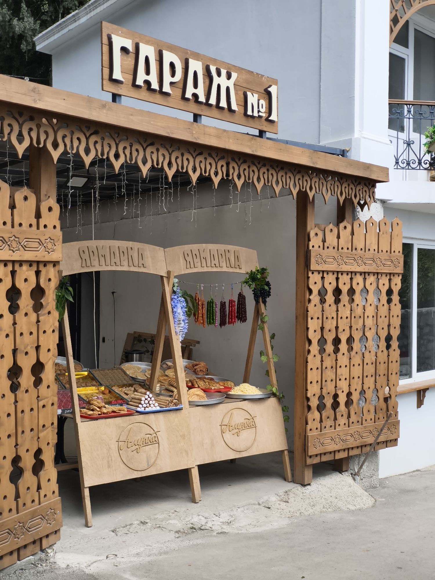 A street-level, eye-level medium shot captures a food stall under a wooden structure with the sign "ГАРАЖ №1" above it. The stall is located on a paved street in what appears to be the city of Unknown, Unknown.
The stall itself is made of light-colored wood and features the word "ЯРМАРКА" on top, written in a Cyrillic script. The front of the stall has the same word, "ЯРМАРКА", inscribed in a circular design. Various trays and dishes filled with food items are displayed on the stall. There is dried fruit hanging on the corner.
To the left and right of the stall are ornately carved wooden panels that function as side walls. These panels have geometric and abstract designs cut into them.
The stall is situated against a light gray wall. There is a white building visible to the right, with a window and what appears to be a balcony on an upper floor.
Above the stall, the wooden structure has a decorative, scalloped edge. Thin strings of lights are suspended from the top of the structure. The lighting is natural, suggesting an outdoor setting.
The image conveys a sense of a small, local food market operating on a city street.