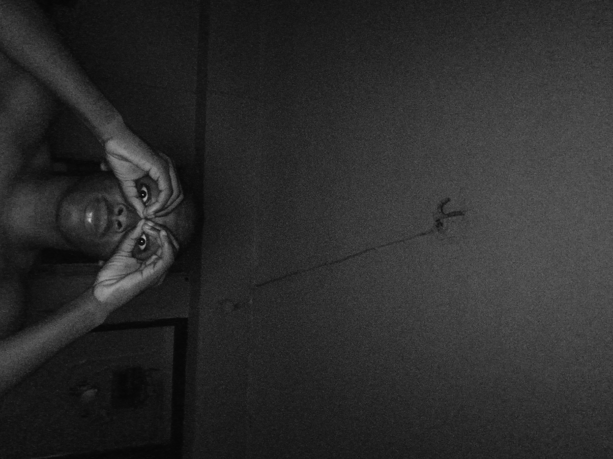 A shirtless individual, possibly male, is positioned on the left side of the frame, facing the camera. Their hands are raised, fingers forming circles around their eyes. The individual appears to be inside a room, with dark walls. On the right side of the image, a cable extends from a wall outlet. The image is in black and white. The location and specifics regarding Lagos, Nigeria are not evident from the provided image.