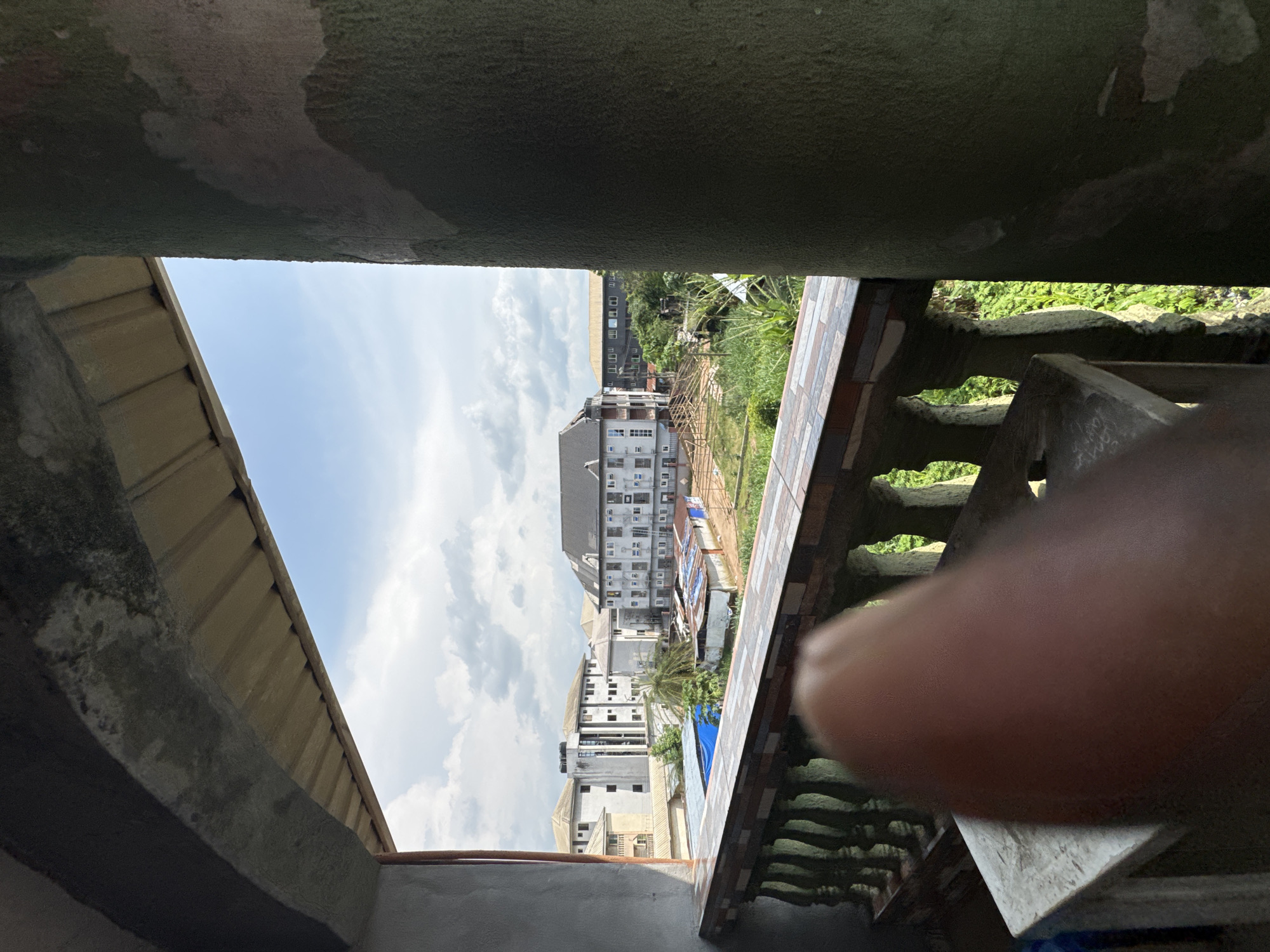 The image shows a view from a balcony overlooking a residential area in Ihiagwa, Nigeria. In the foreground, a blurry thumb obscures part of the view. A concrete balustrade with decorative tiles runs along the edge of the balcony. Beyond the balcony, several multi-story buildings are visible, some appearing to be under construction with exposed wooden framing. Lush green vegetation surrounds the buildings. The sky is partly cloudy. The scene depicts a typical urban landscape in Ihiagwa, characterized by its residential buildings and ongoing development.