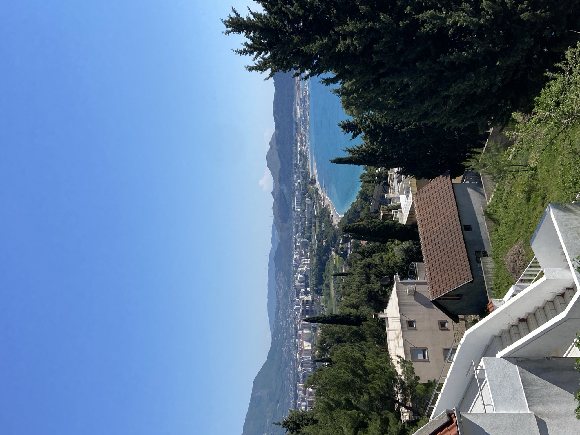 This is a scenic, high-angle view of Šušanj, Montenegro, showing a coastline with a beach and buildings nestled between the mountains and the Adriatic Sea. A clear blue sky dominates the upper left. The landscape includes residential buildings with varying architectural styles, with tile roofs and white walls. Lush green trees and vegetation surround the buildings, adding to the landscape. A set of white stairs leading down the hillside is visible in the foreground.