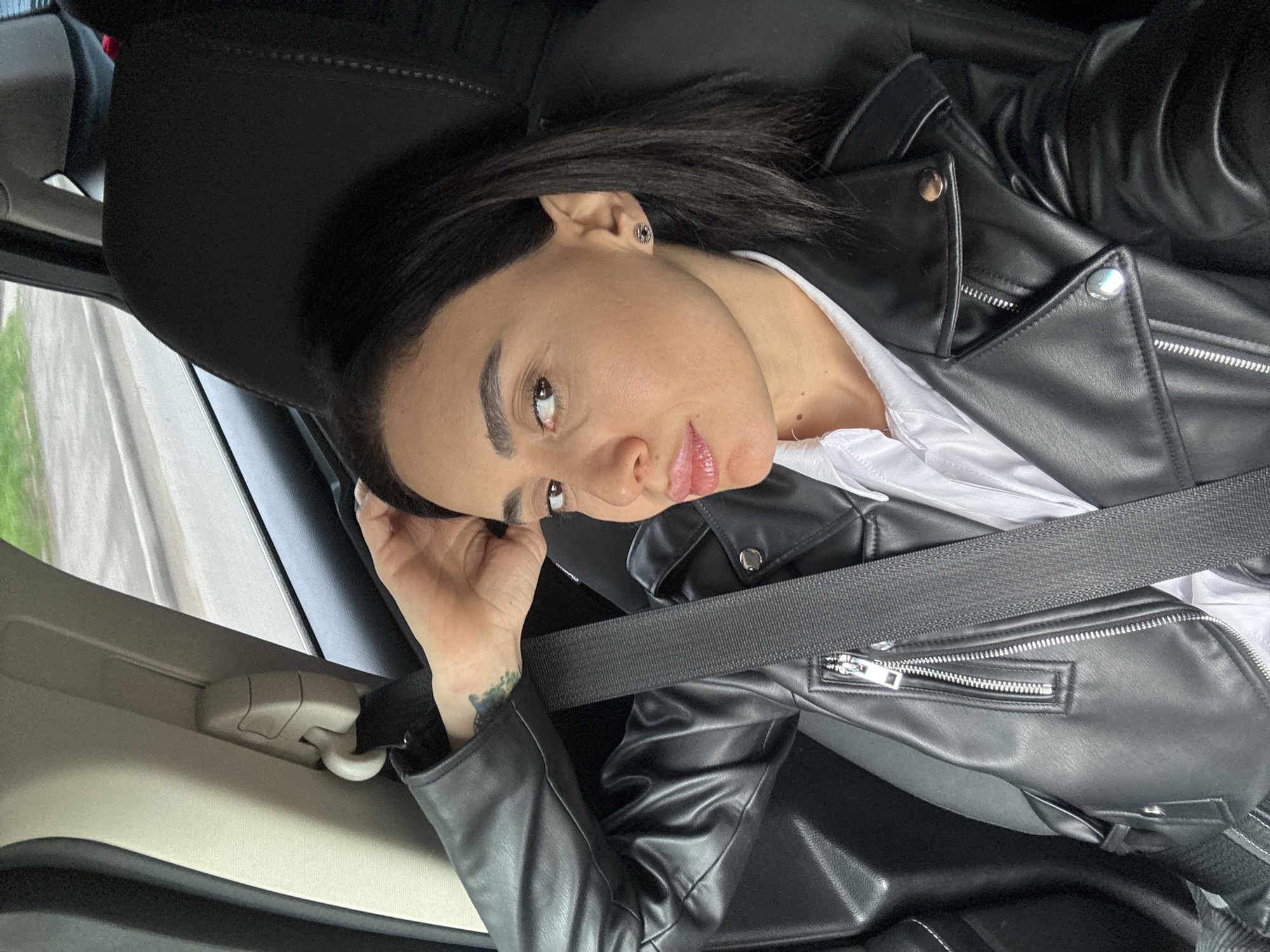 A woman is seated in a car, wearing a black leather jacket over a white shirt and a seatbelt. The car is in motion, as evidenced by the blurred background seen through the window. The woman is looking at the camera with a slightly tilted head and a hand near her face. The location appears to be a city street, possibly in Tbilisi, Georgia, as it includes standard road infrastructure.