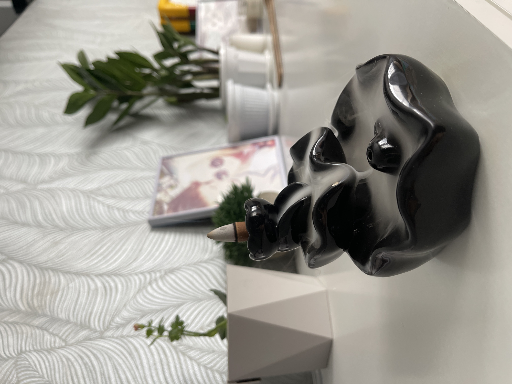 The image captures a close-up, slightly rotated view of objects arranged on a surface, likely a bed or a table covered with a textured, white fabric. The focal point is a black, glossy incense burner with an incense stick inserted into it. The incense stick appears to be lit, and smoke is emanating from it.
Additional objects are present in the background, including a green leafy plant, a postcard or photo with a blurred image, and several small white containers. A white geometric-shaped container is also visible.
The image provides no information to suggest the location is in Izhevsk, Russia.