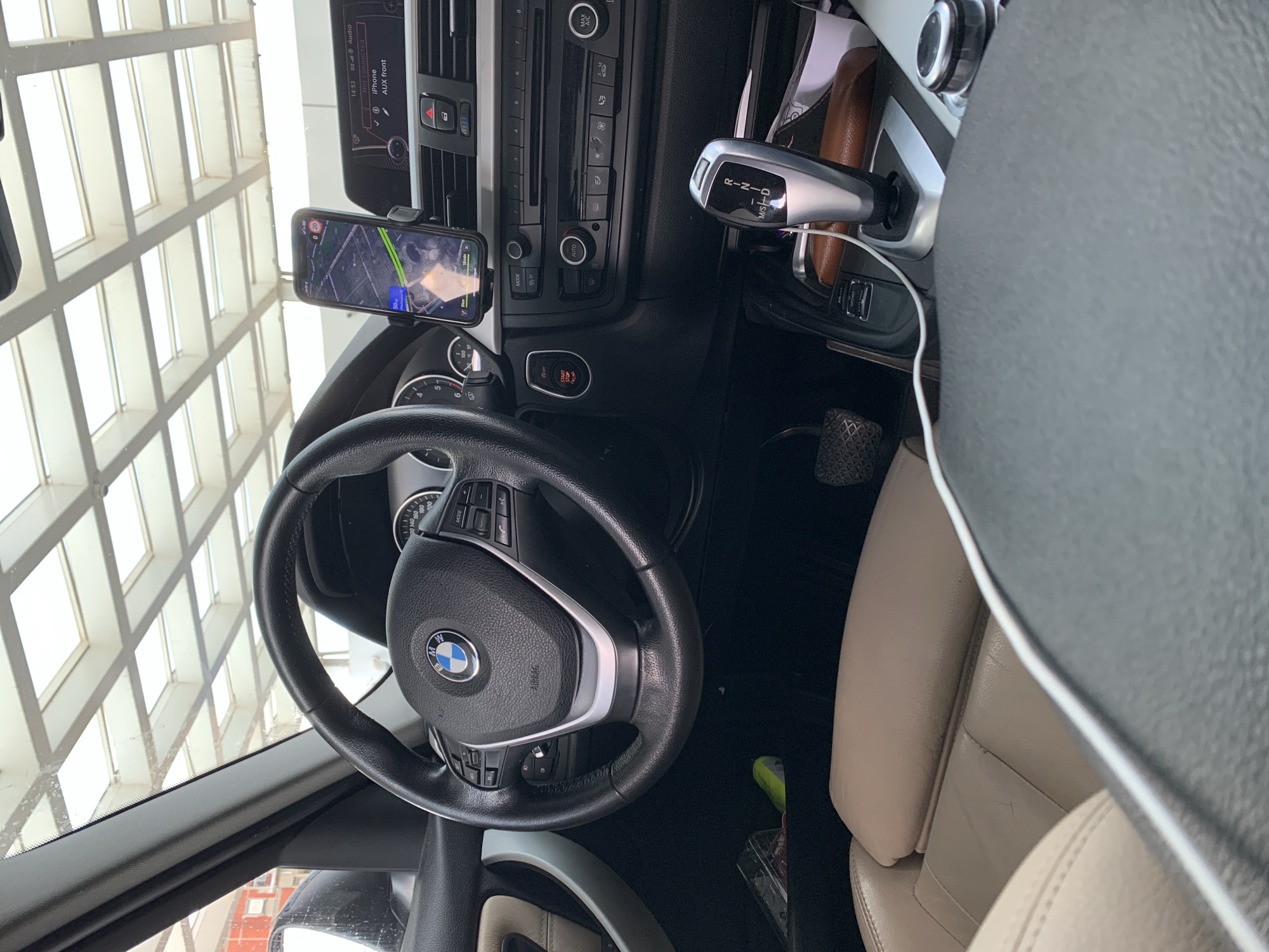The image shows the interior of a BMW car, specifically the driver's side. The focus is on the steering wheel, dashboard, and center console. A smartphone is mounted on the dashboard, displaying a map. The gear selector is visible, along with the car's infotainment screen. The seat is upholstered in tan leather. The car is likely parked indoors.
Due to the visible map on the smartphone, it is possible the car is located in the city of Vozdovac Urban Municipality, Serbia.