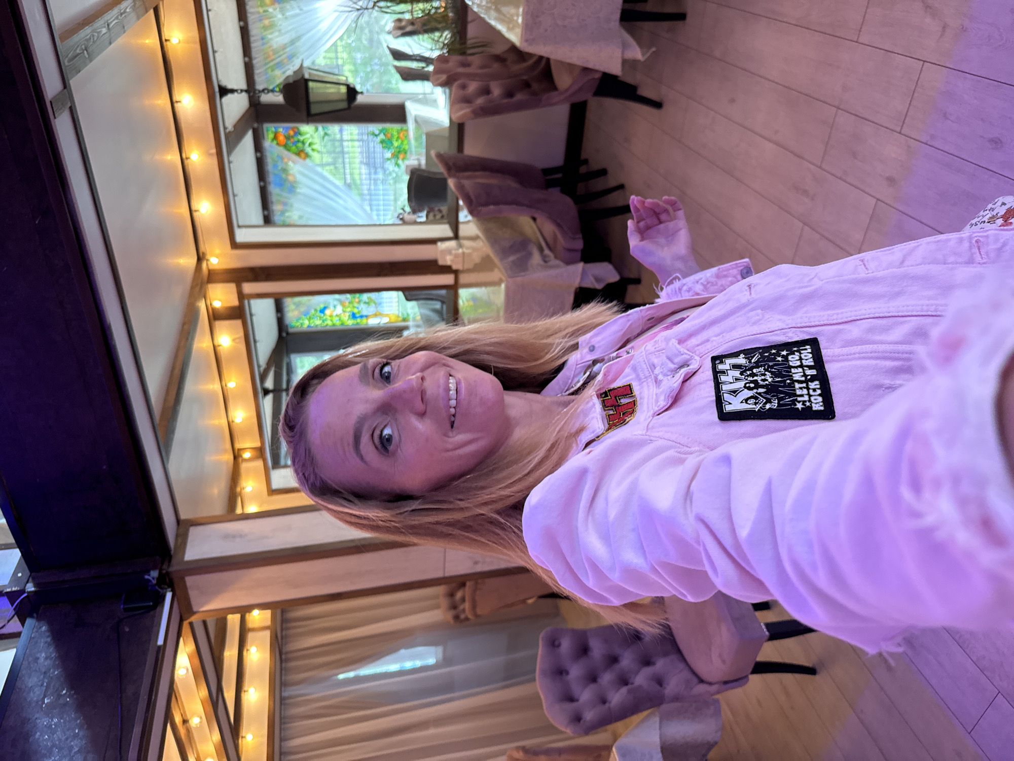 The media file shows a woman taking a selfie. She is wearing a pink denim jacket with a patch on the left breast pocket that says "KISS LET ME GO ROCK 'N' ROLL". The woman has blonde hair and is smiling at the camera. She appears to be indoors, possibly in a restaurant or event space, with wooden floors and decorative string lights. Stained glass windows with colorful patterns are visible in the background, along with upholstered chairs and tables. The lighting in the room is warm, suggesting it might be evening or a dimly lit interior. The woman's facial expression conveys happiness and confidence. The setting appears to be in Lomonosov, Russia, based on the provided location context.