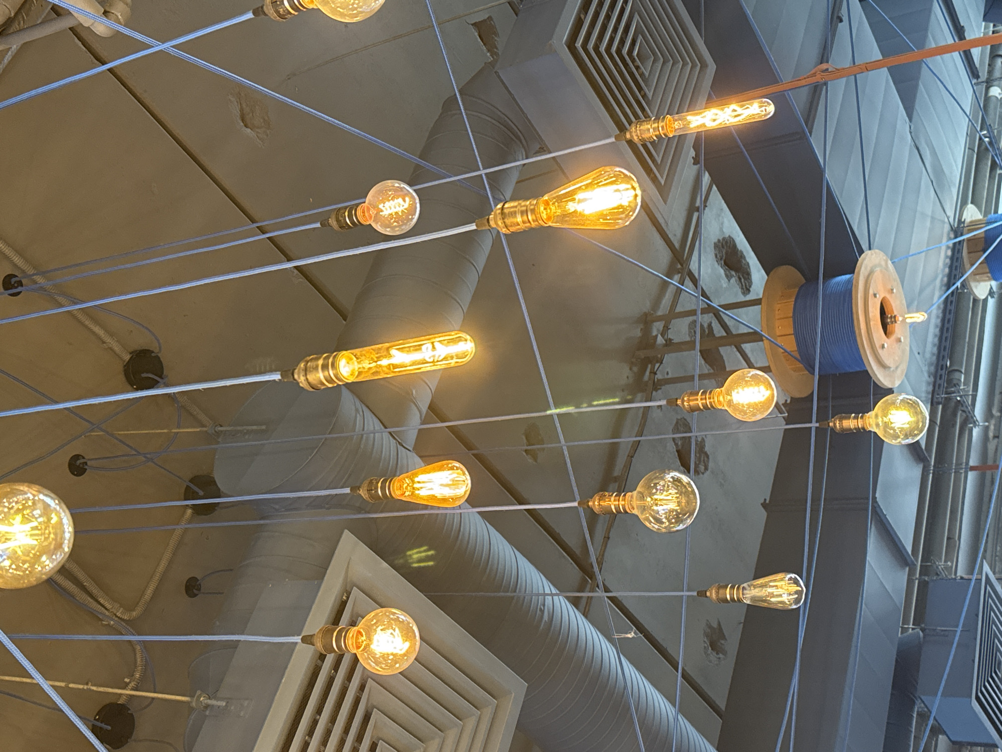 This image, taken from an upward perspective in an indoor setting in Saint Petersburg, Russia, showcases an industrial-chic ceiling design. The primary subjects are numerous exposed Edison-style light bulbs, each with glowing filaments, suspended at various heights and angles. These bulbs emit a warm, amber light and are interconnected by a network of thin, light blue cables that crisscross the frame, forming a dynamic, web-like pattern against the ceiling.
The background reveals an exposed ceiling structure featuring large, circular metallic air ducts, some running horizontally and others vertically, alongside square and rectangular ventilation grates. A textured concrete or plaster surface is also visible. Adding to the industrial aesthetic, two large wooden spools, wound with bright blue electrical cable, are prominently displayed on the right side of the frame, serving as both functional and decorative elements.
No people or specific activities are visible within the frame. The warm artificial lighting suggests an evening ambiance or a deliberate design choice to create a cozy or modern atmosphere. There is no discernible text in the image.