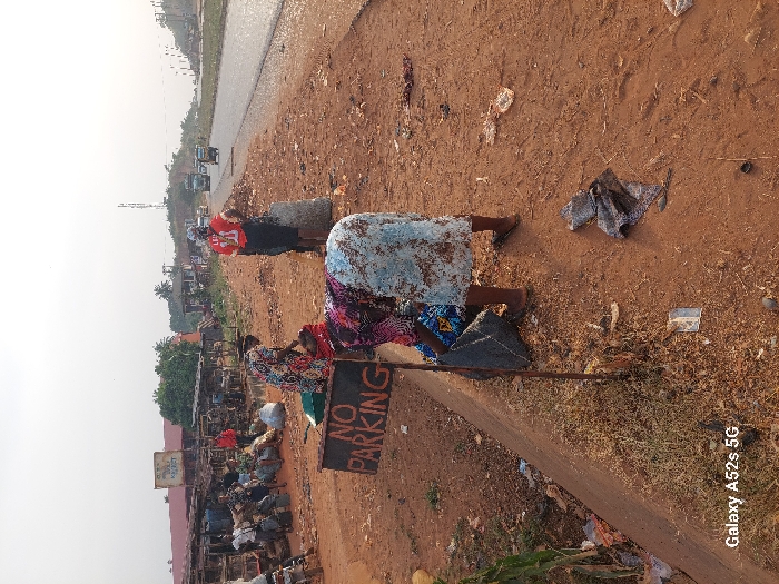 The image depicts a roadside scene in Awkuzu, Nigeria, characterized by market activity and ground-level commerce. In the foreground, a crudely constructed "NO PARKING" sign on a metal pole is prominent on the left. The ground is reddish-brown dirt, dry and sparsely vegetated, with scattered litter including fabric scraps, plastic, and paper.
Three women are central to the scene's midground. One woman, in the immediate foreground, wears a patterned long-sleeved top and a light blue and brown patterned skirt, bending low to sort or pack items into a dark grey sack. Her feet are visible in dark flip-flops. Slightly behind her, another woman in a floral dress is similarly crouched, near a green basin or container, engaged in a packing activity. Further right, a third woman stands upright, her back to the viewer, wearing a red jersey with the number "11" and dark bottoms, positioned next to a large, light-colored sack filled with contents.
To the left, a series of open-air market stalls with wooden frames and corrugated metal roofs are visible, surrounded by numerous large sacks, suggesting a market area. A sign, partially obscured, reads "Paddy Market." Multiple individuals are seated or standing within this market section.
To the right, a two-lane paved road extends into the distance. Vehicles, including a large green truck and two motorized tricycles ("Keke Napep"), are observed moving away from the camera on the road. The shoulder of the road is unpaved and consists of reddish-brown dirt. In the distant background, a tall metal communication tower stands against a hazy, pale sky, with scattered green foliage visible across the landscape. The overall lighting suggests an outdoor environment, possibly under a bright or slightly overcast sky.