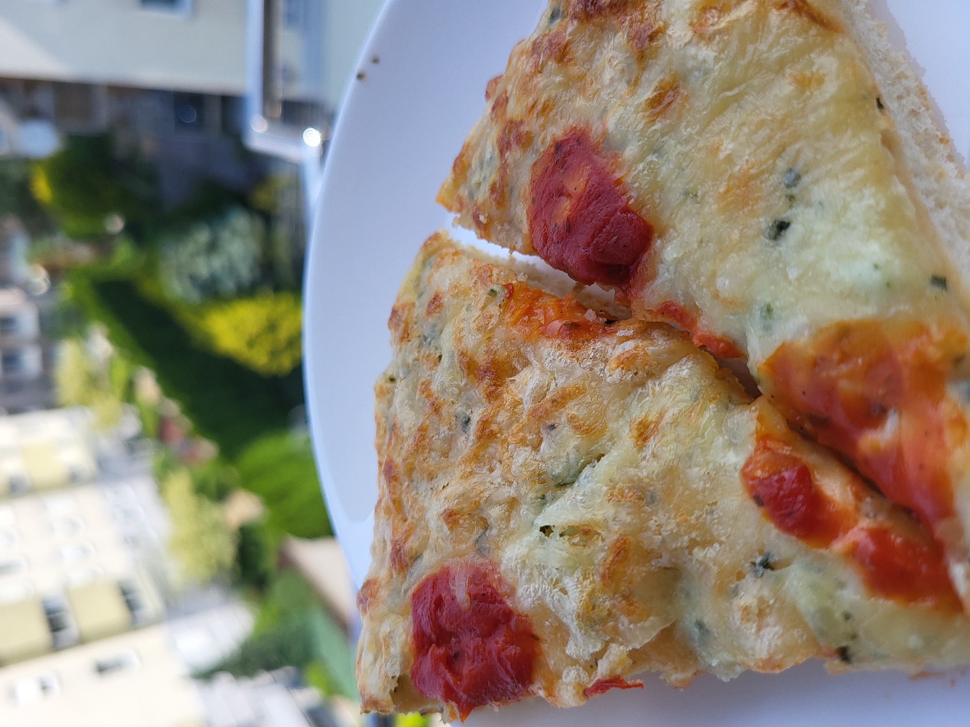 Two slices of pizza with red sauce and melted cheese are on a white plate. In the background, blurred buildings and greenery are visible. The location is unknown.