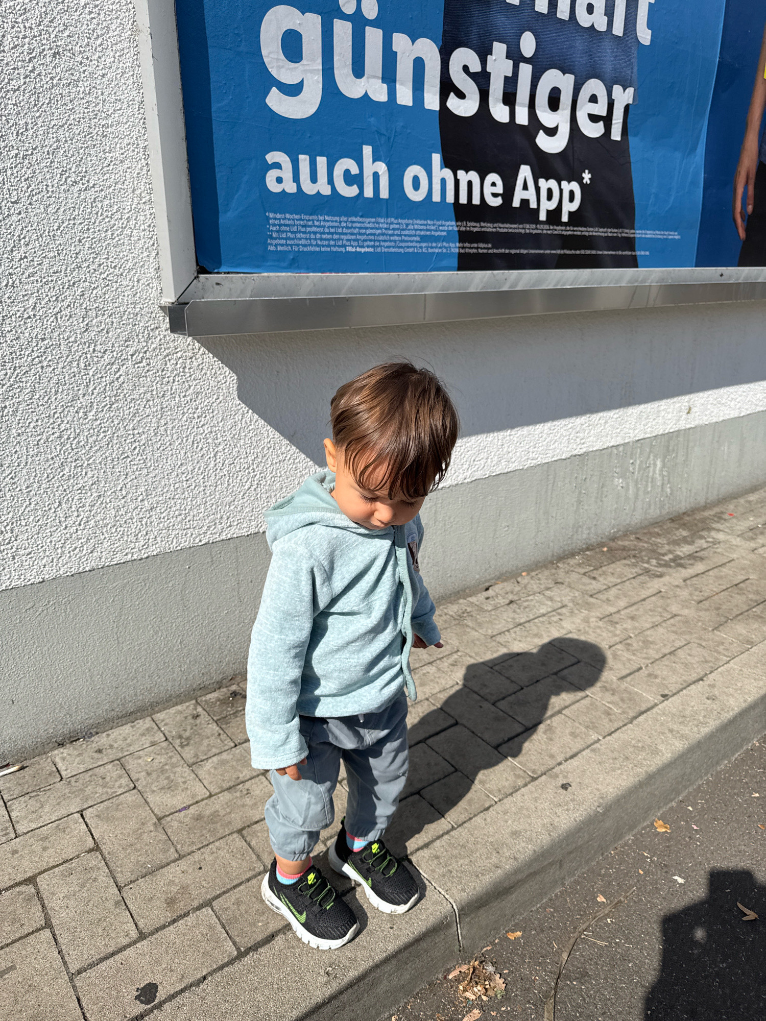 In Überherrn, Germany, a young child stands on a sidewalk next to a white textured wall. The child is wearing a light blue hooded sweatshirt, gray pants, and black and neon green sneakers with blue and pink striped socks. The child is looking down towards the ground and is casting a shadow on the sidewalk. A large blue banner with white text hangs on the wall behind the child. The sidewalk is made of concrete pavers. The ground next to the sidewalk is asphalt with some dry leaves scattered on it. The sun is casting strong shadows, indicating it is daytime.
