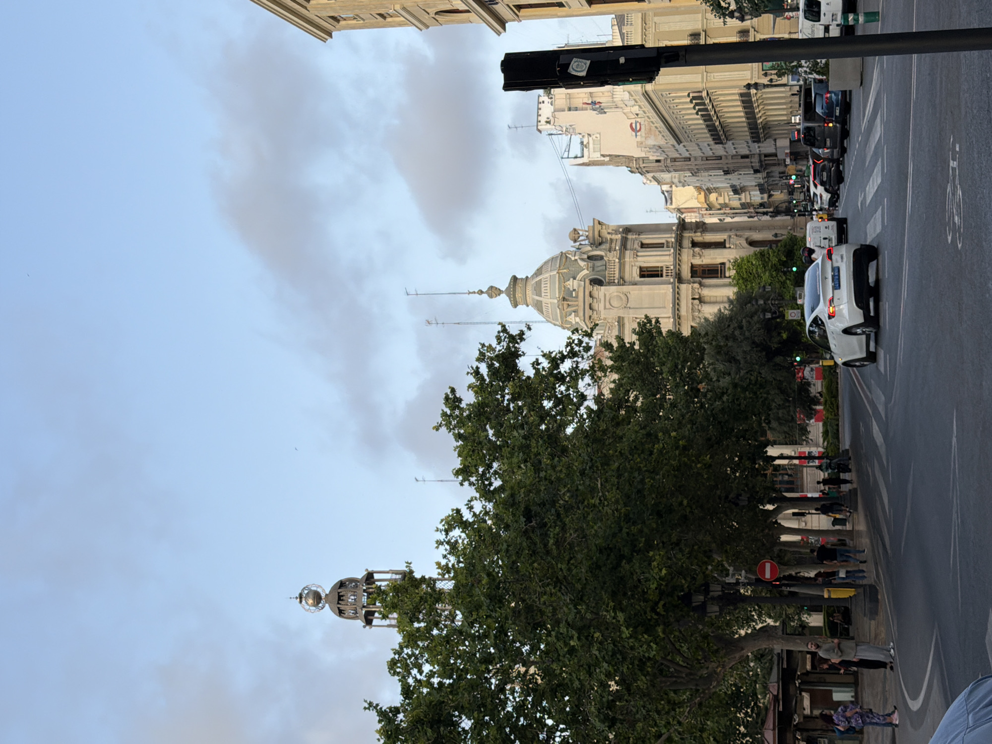 The image depicts a street scene in a city, likely in Europe given the architecture. A white Tesla is visible at a crosswalk, as are several other vehicles, including a van and a blue car. Traffic lights are present, with some showing green. People are walking on the sidewalk and crossing the street. In the background, there are ornate buildings with detailed facades and domed roofs, as well as trees lining the street. A tall, decorative tower with a spherical top is also visible. The sky is partly cloudy.