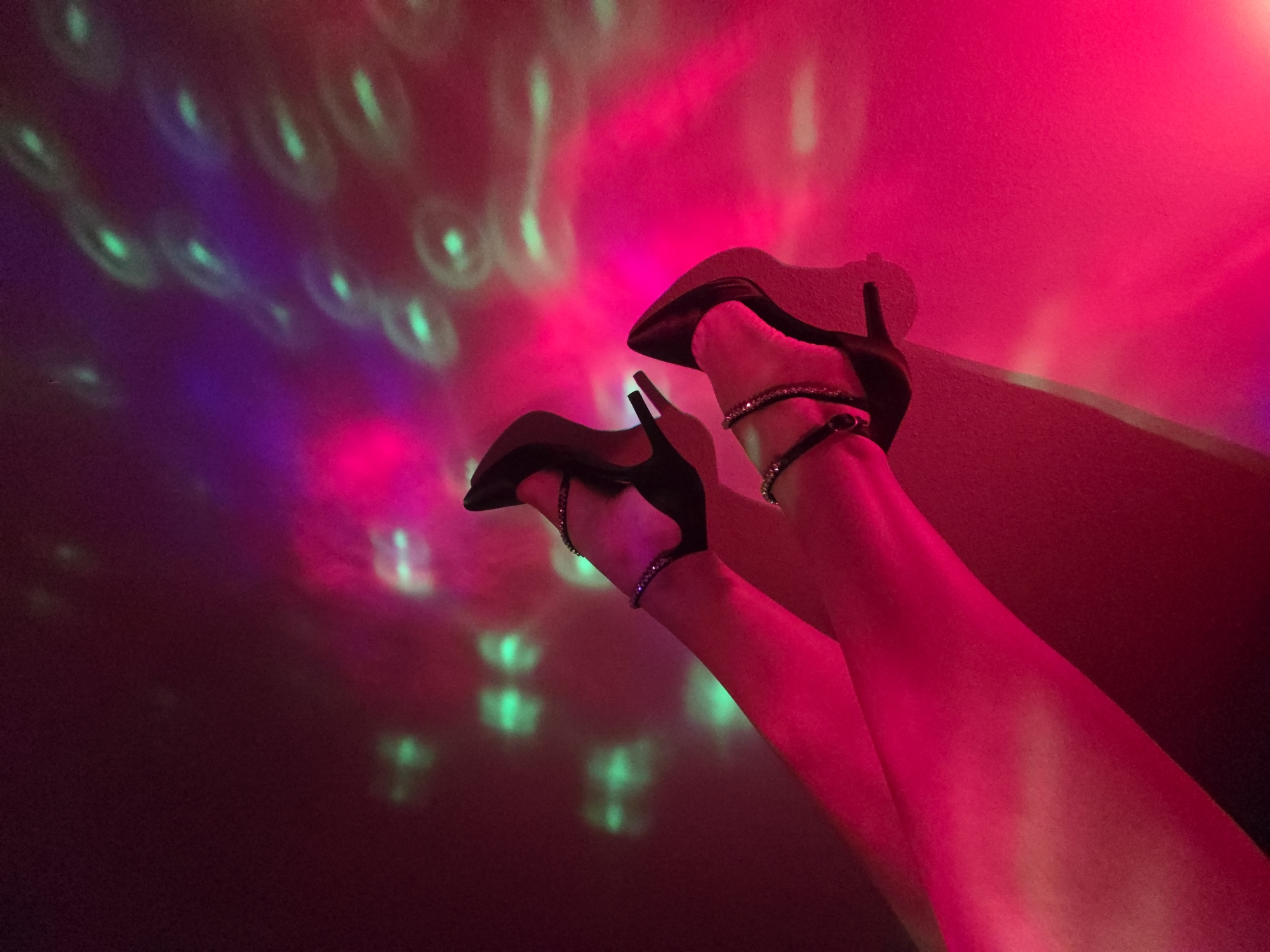 In an unidentified city, an interior space is illuminated by multi-colored lights, predominantly pink, red, and purple, with green circular patterns scattered across the background. The scene features a pair of legs, positioned vertically, with black high-heeled shoes on the feet. The shoes have thin straps around the ankles. The lighting casts shadows and highlights on the legs.