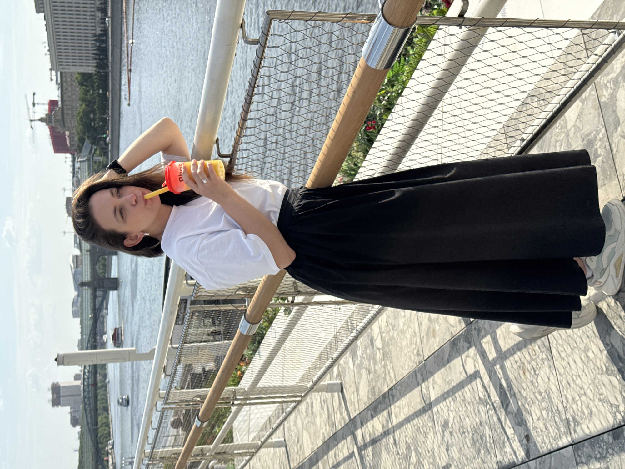 A woman is drinking from a cup while standing on a bridge. She is wearing a white shirt, a black skirt, and white sneakers. She has brown hair and is wearing a watch on her left wrist and a ring on her left hand. She is holding the cup with her right hand and has her left hand on her head. The cup is yellow, orange, and red and has a straw. The bridge has a metal railing and a wooden handrail. Behind the woman is a river with buildings on the other side. The sky is blue and there are some clouds.